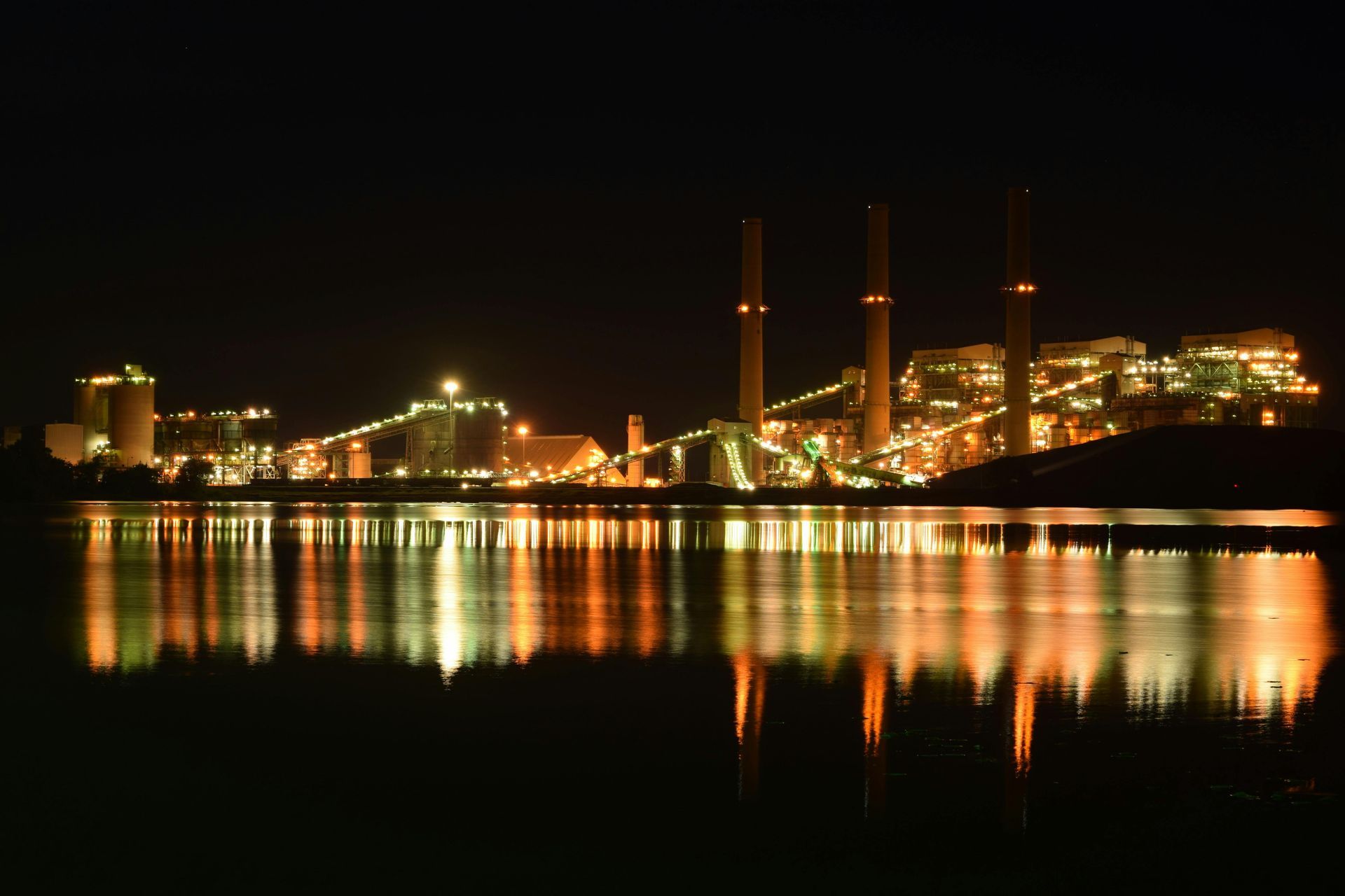 Industrial complex at night, reflecting lights on water. Three smokestacks rise above.