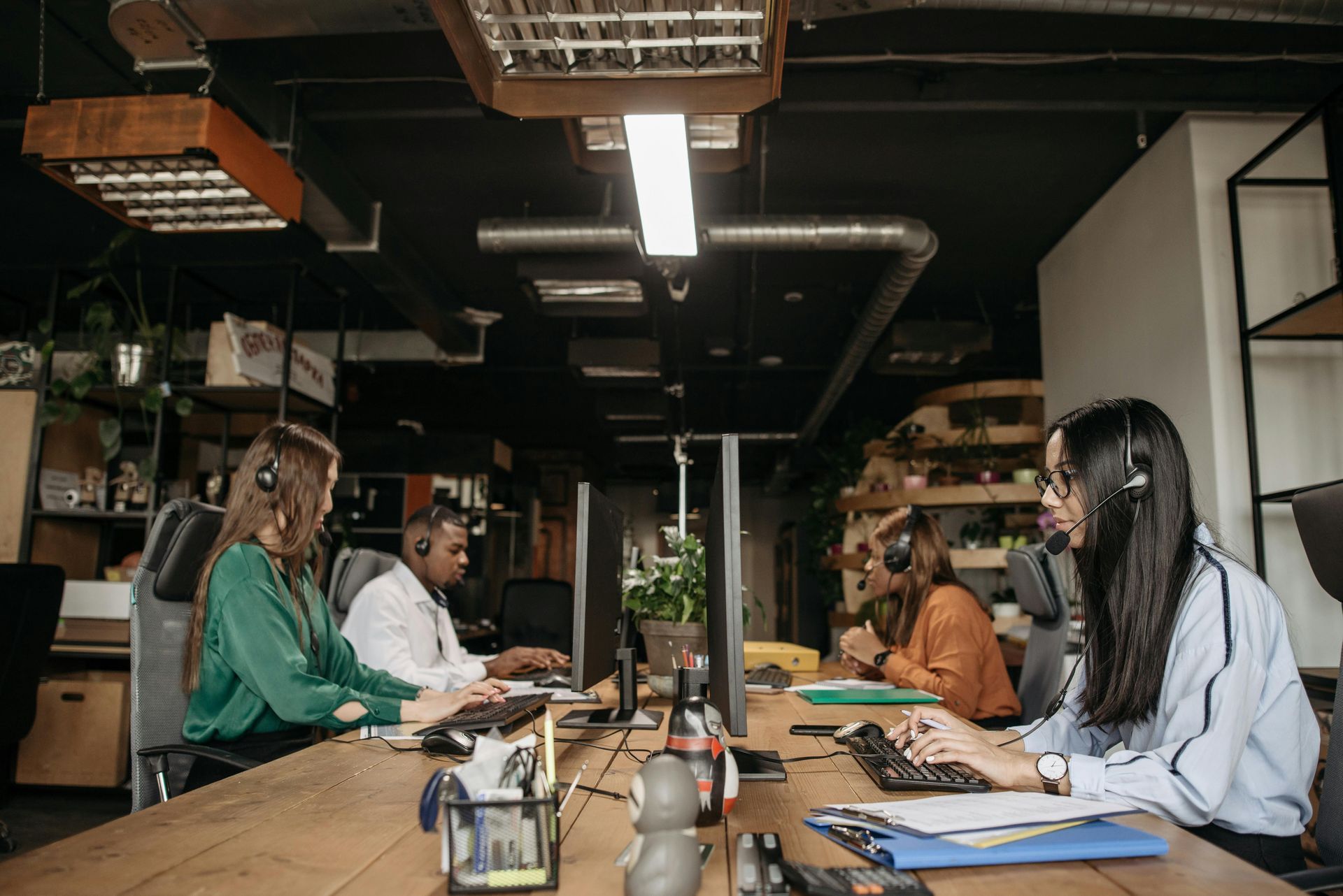 Office workers with headsets typing at computers in a row. Dark ceiling, wooden desk.