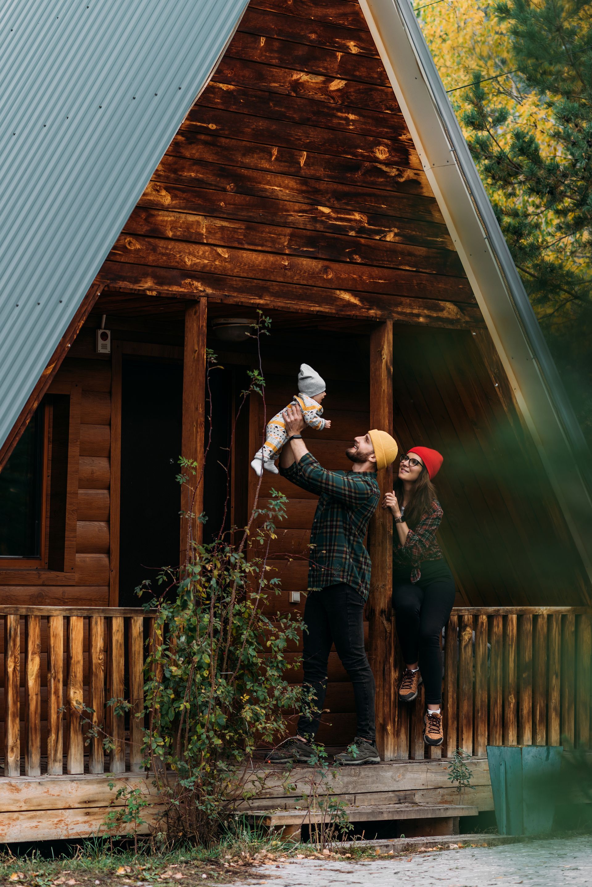 Family outside a log cabin; father holding up a baby; mother watches.