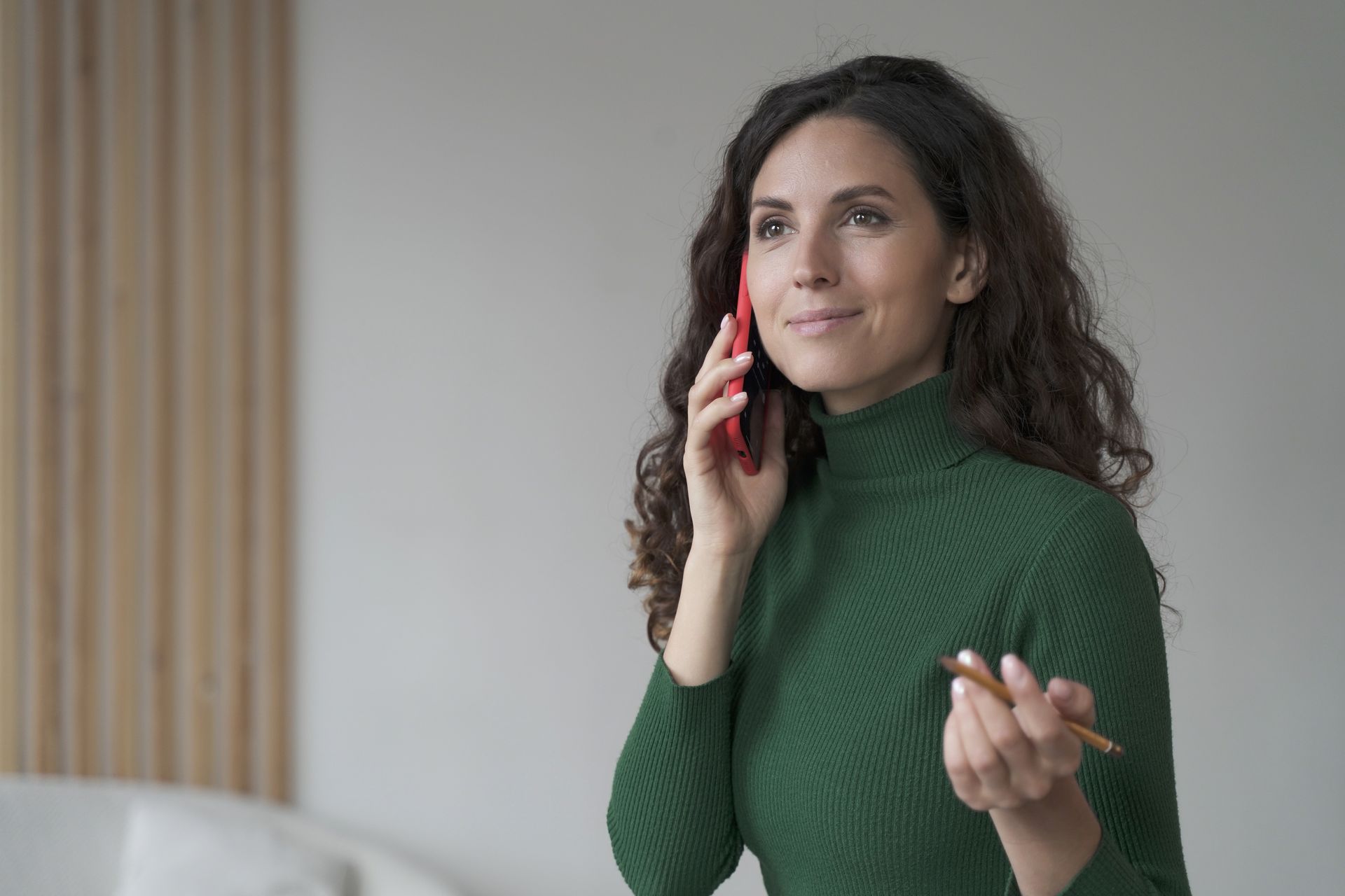 Woman with curly hair in a green turtleneck smiles while talking on a red phone, holding a pen.