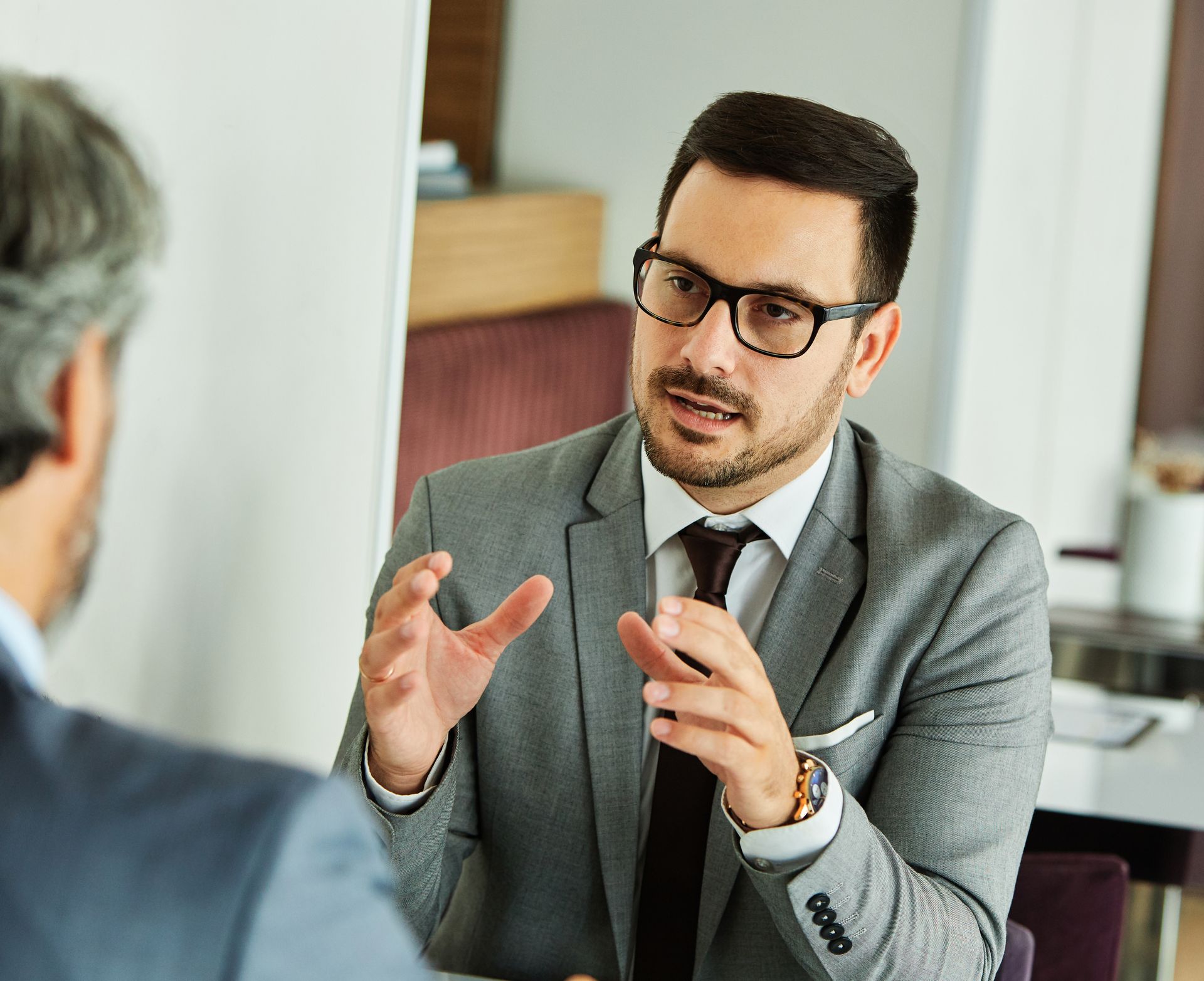 Man in suit with glasses gesturing, speaking to another person at a table.