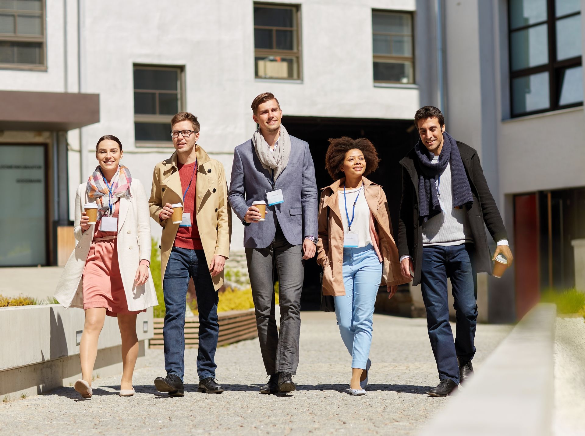 Five people walking outside a building, holding coffee cups and wearing scarves.