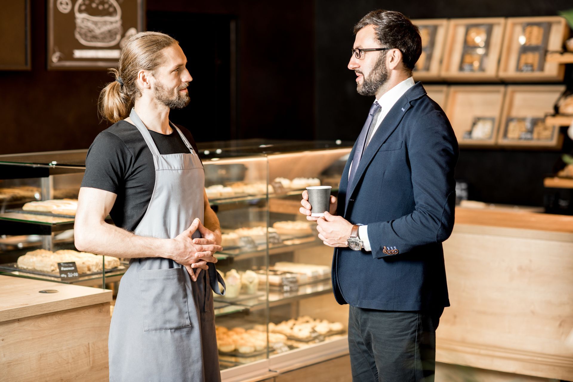 Barista and suited man talking in a bakery, man holding a coffee cup, pastries in the display case.