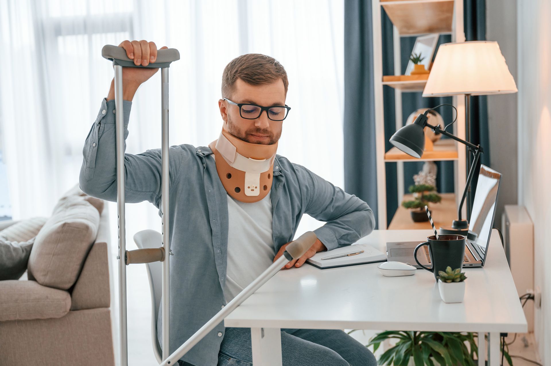 Man in neck brace using crutches, working at a desk with a laptop.