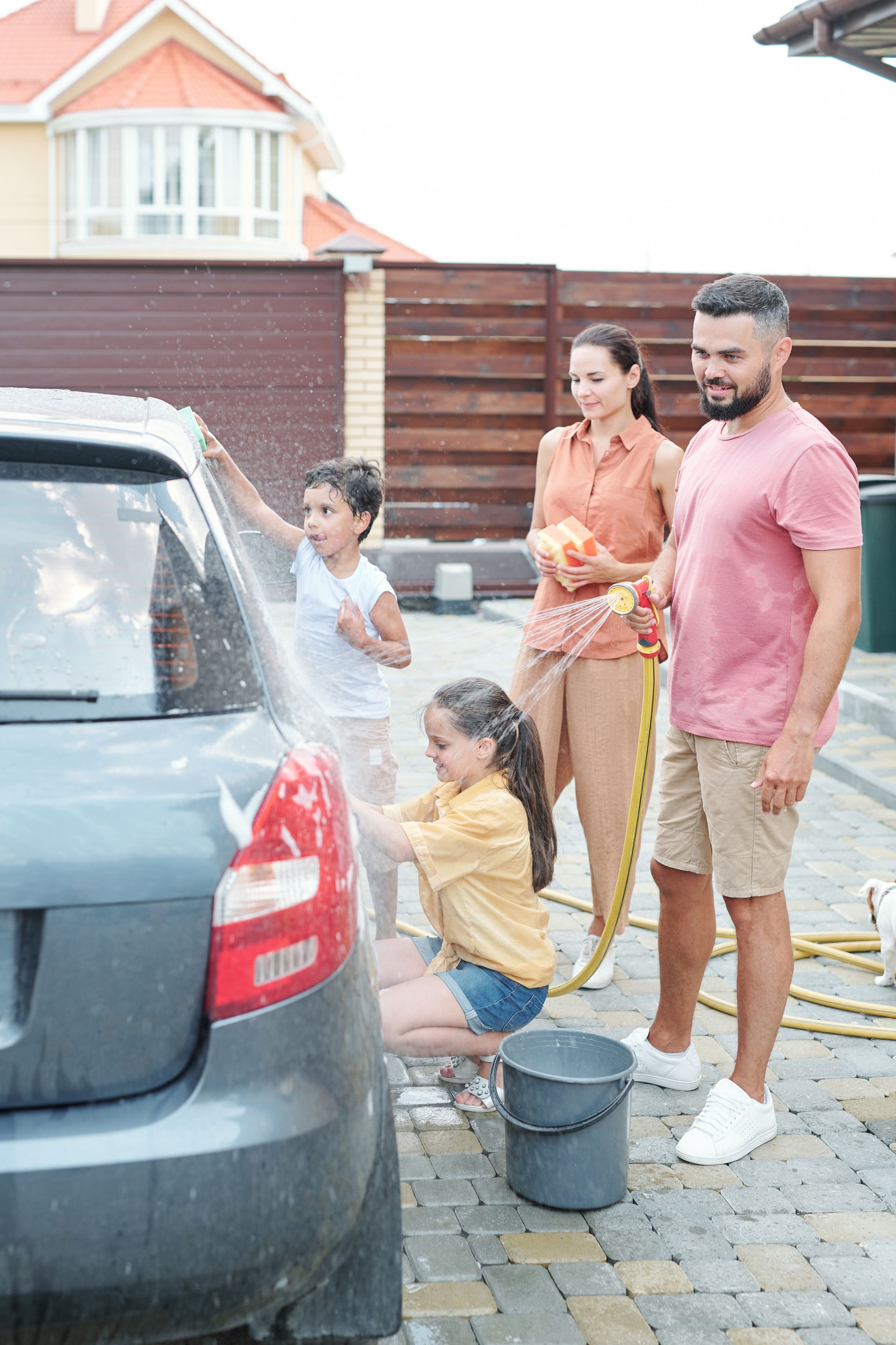 Family washing a gray car in a driveway; children washing with sponges, parents looking on.