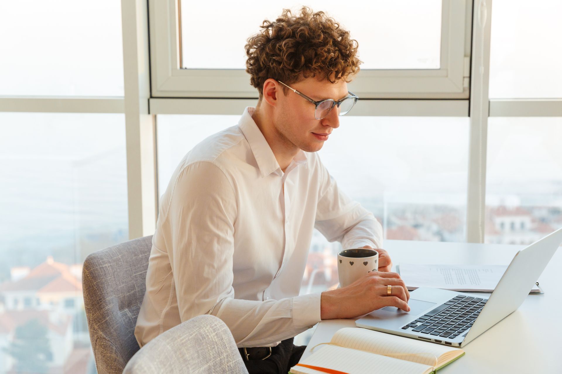 Man with curly hair and glasses works on a laptop at a bright desk, holding a mug.