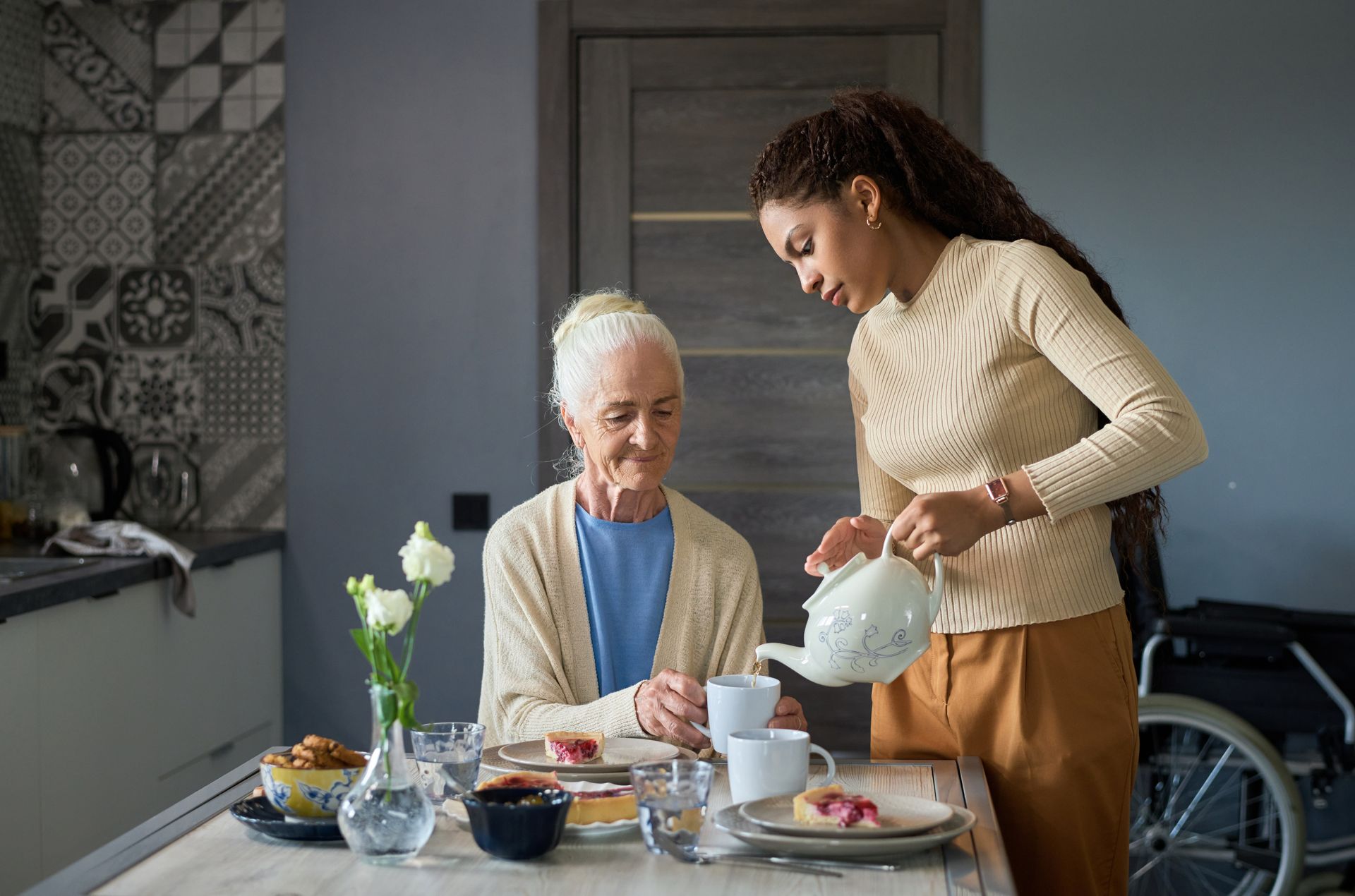 A person pouring tea for another person at a table. Table has food, flowers. Wheelchair visible.