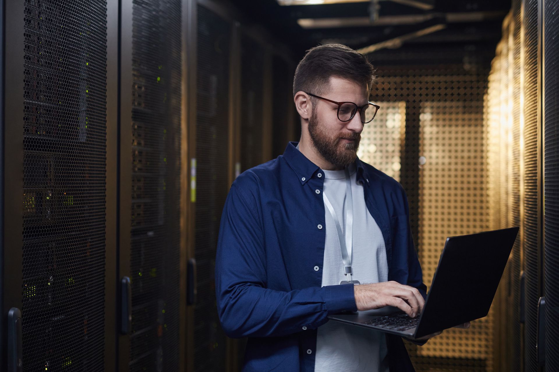 Man in a data center using a laptop, looking at the screen. He wears glasses, a blue shirt, and a lanyard.
