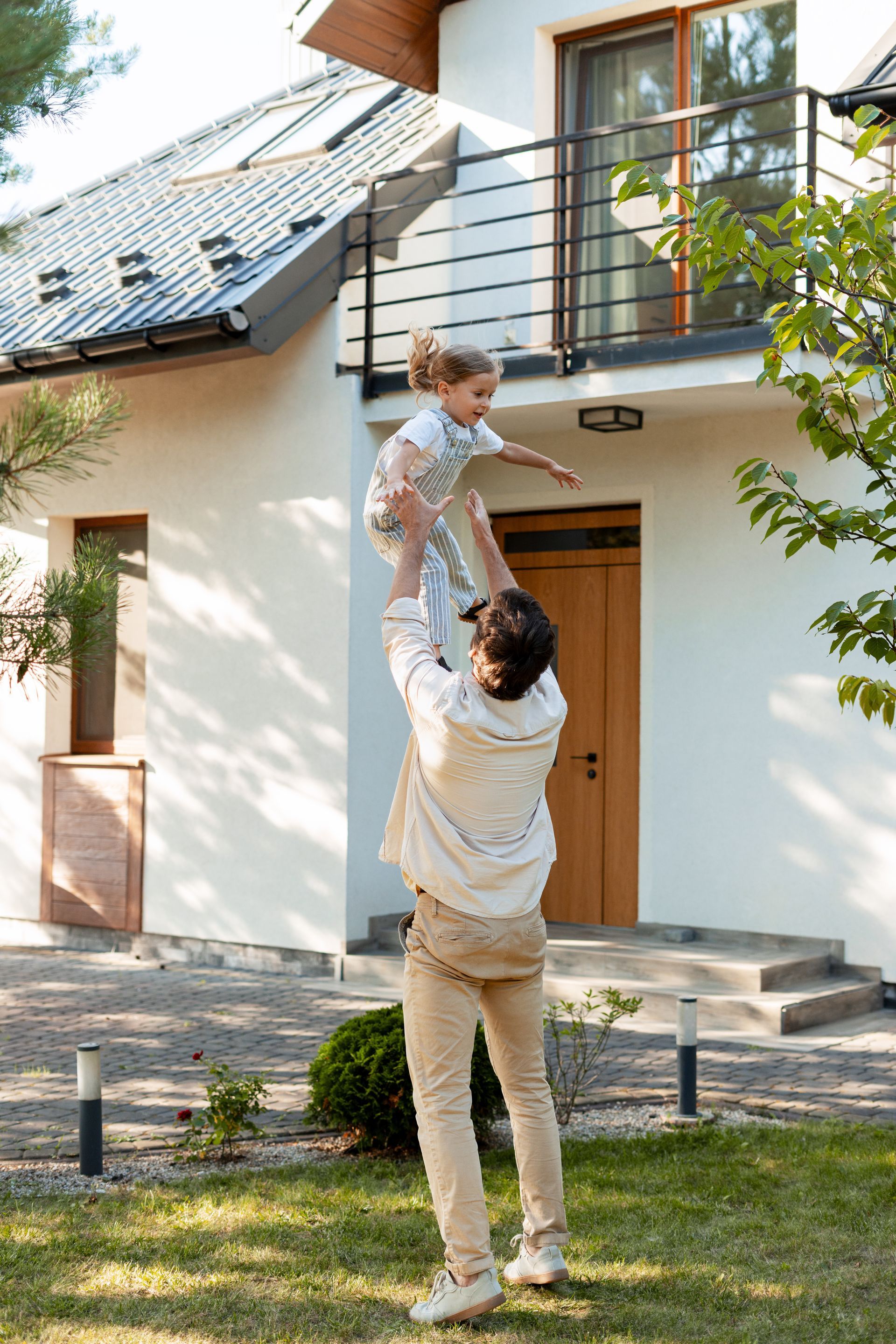 Man throws a child in the air in front of a house with a balcony.