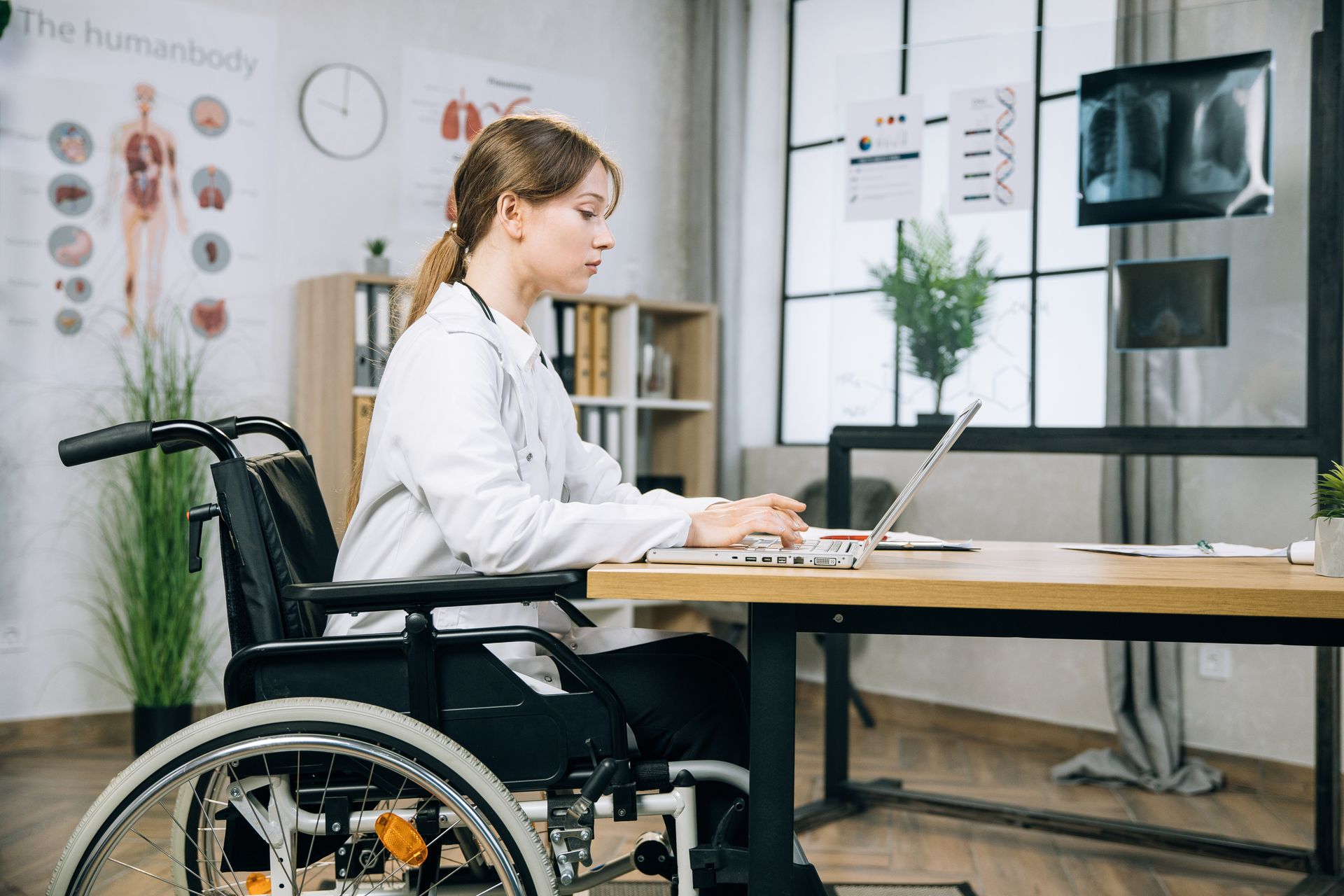 Woman in a wheelchair, wearing a lab coat, using a laptop at a desk in a medical office.