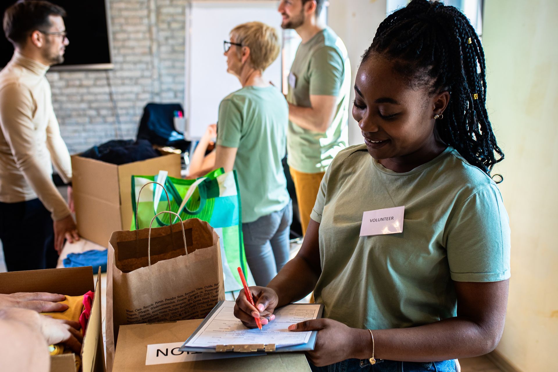 Volunteers sort donations in a room. Woman writes on a clipboard next to cardboard boxes. 