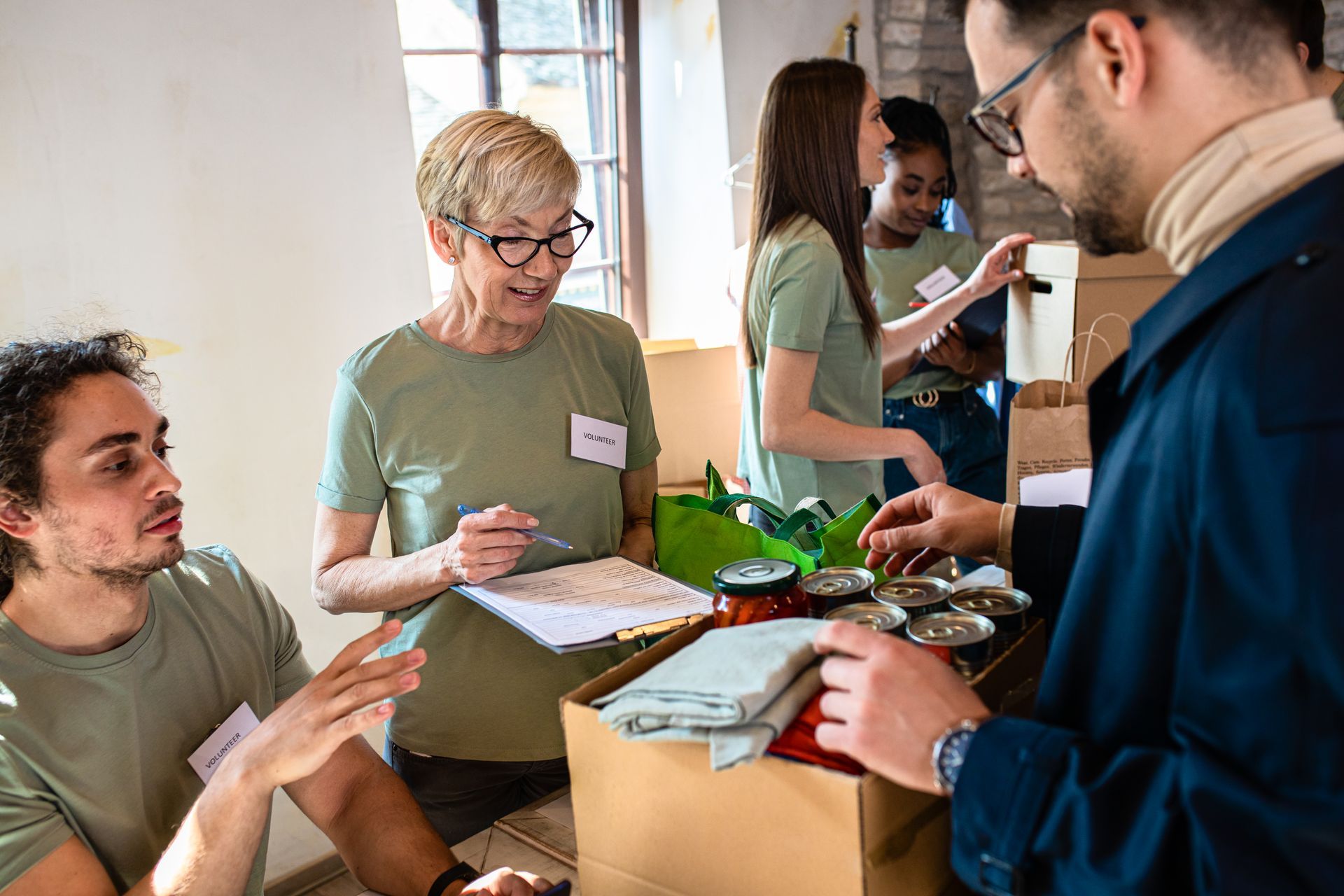 Volunteers packing food items at a distribution center, interacting with people, in a bright room.