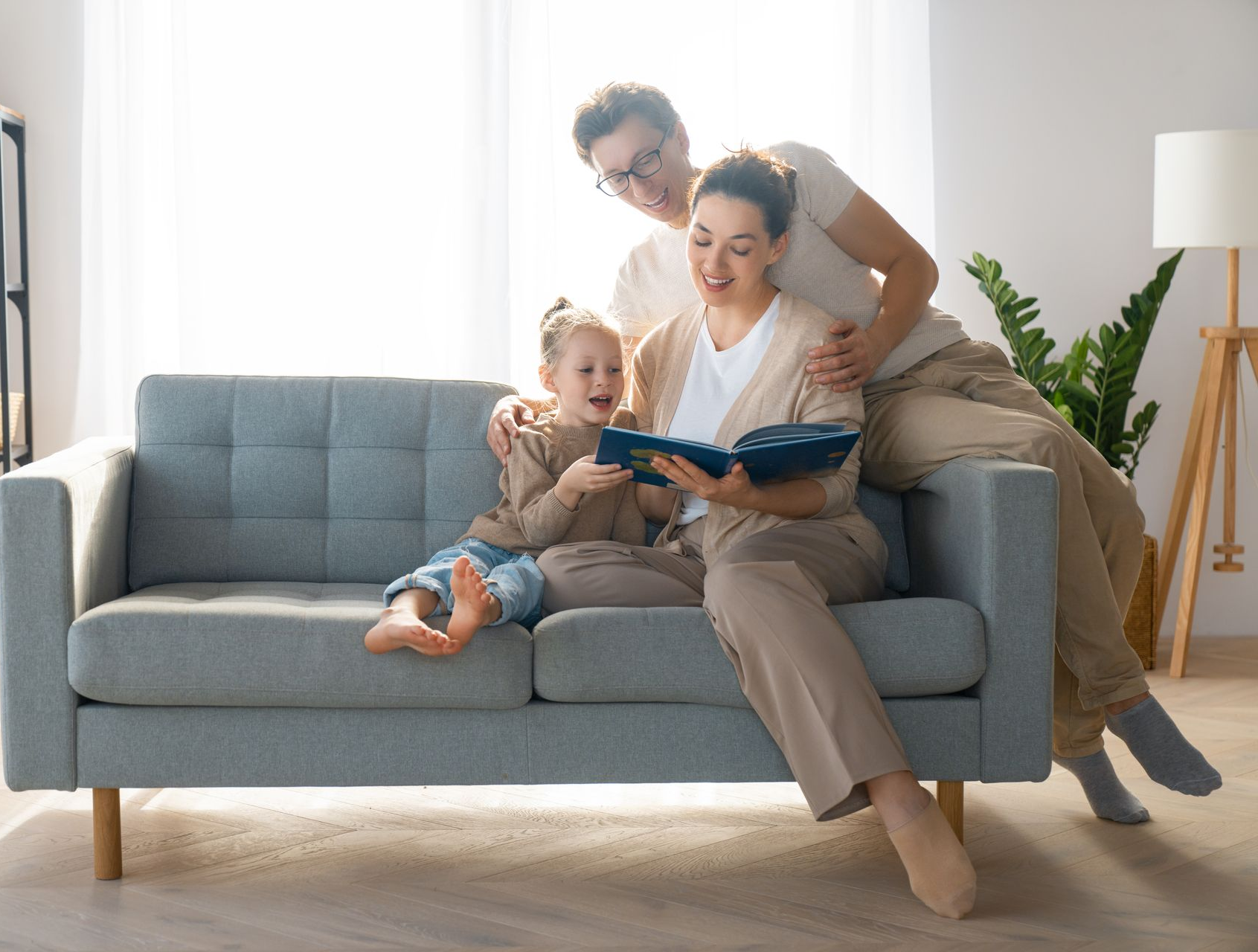 Family reading together on a gray couch: mother reads to child, father looks on, bright living room.