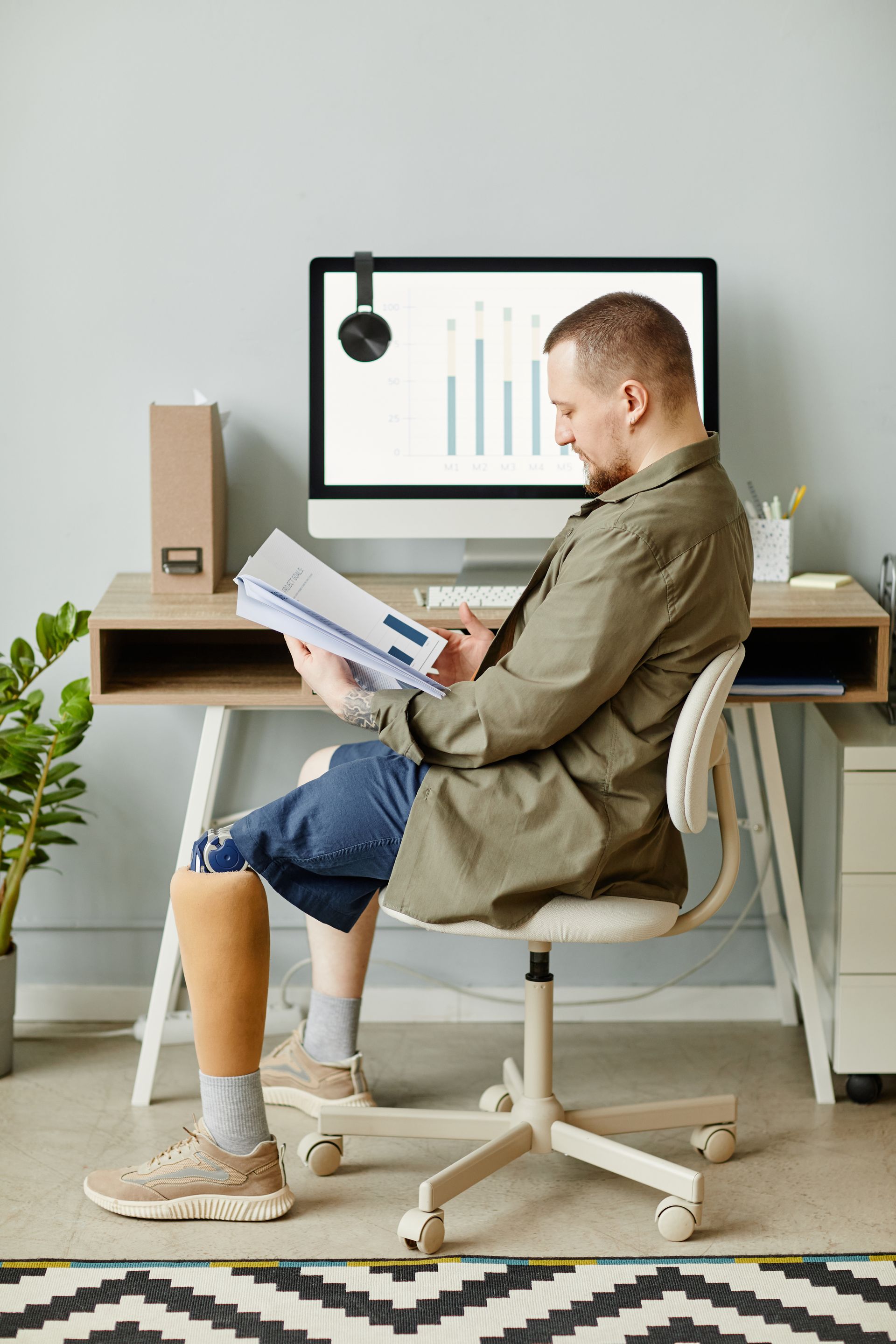 Man seated at desk, reviewing documents, wearing shorts, with prosthetic leg. Computer and office supplies.