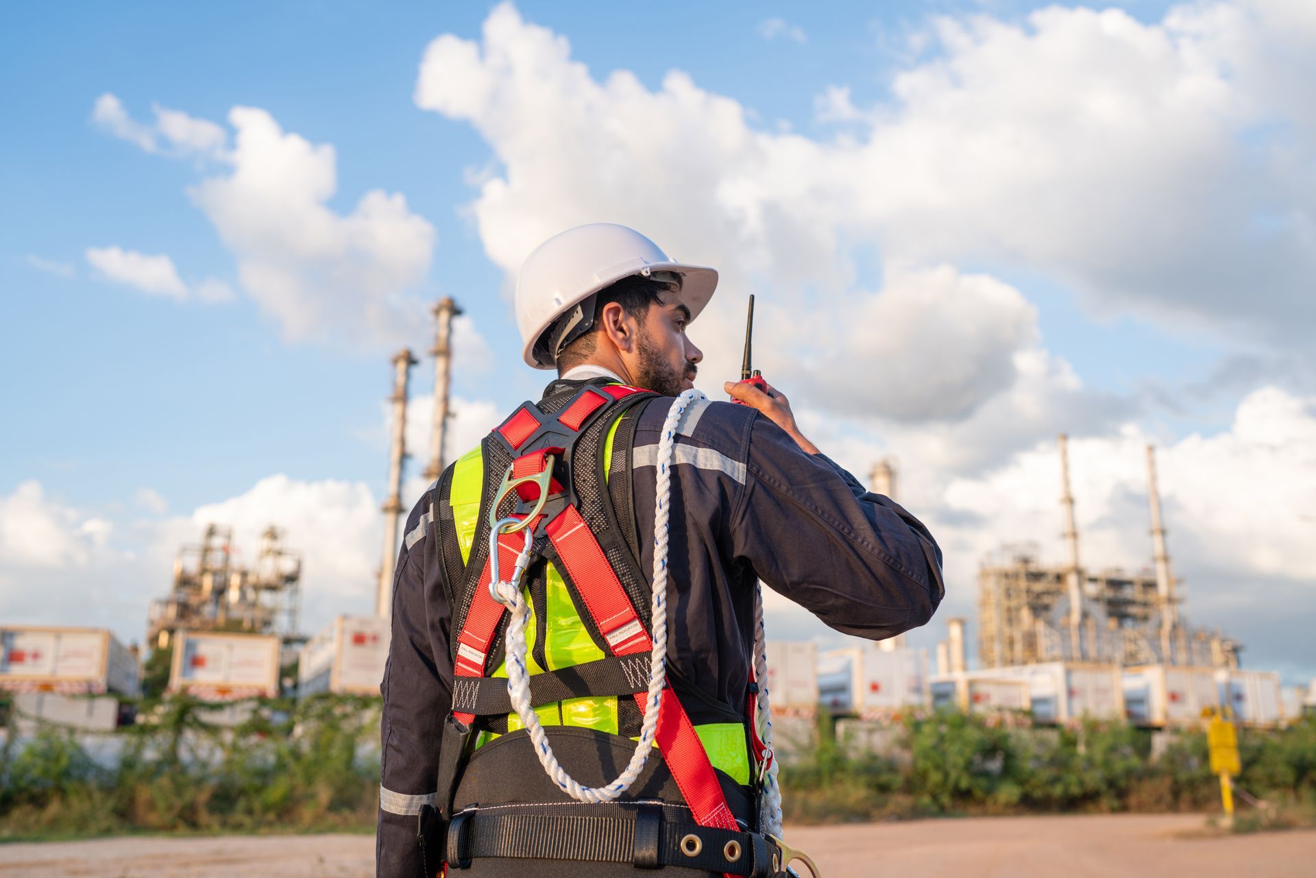 Construction worker in safety harness holding a radio, facing away from the camera, with a factory in the background.