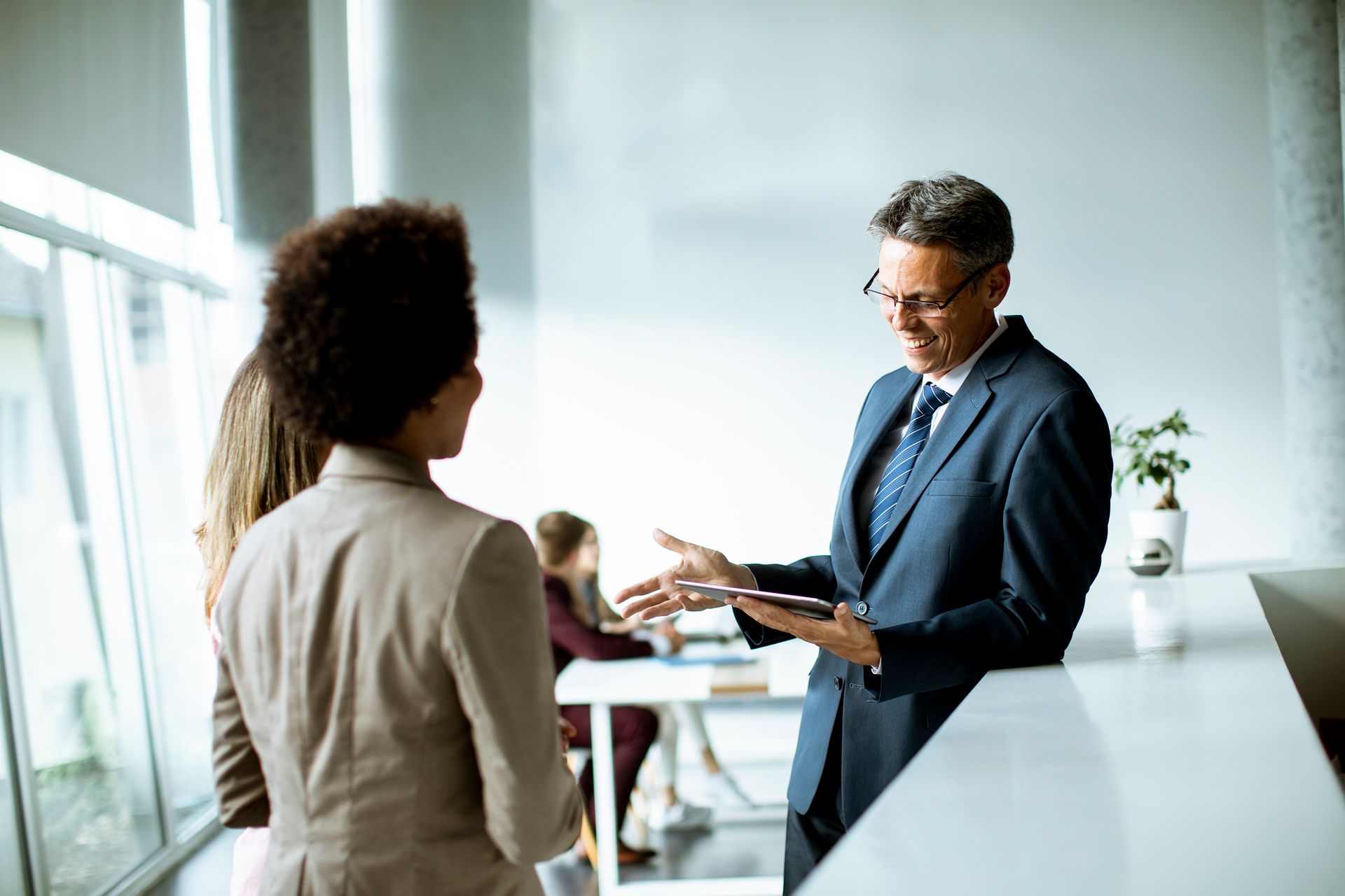 Man in suit gestures towards two colleagues in an office setting.