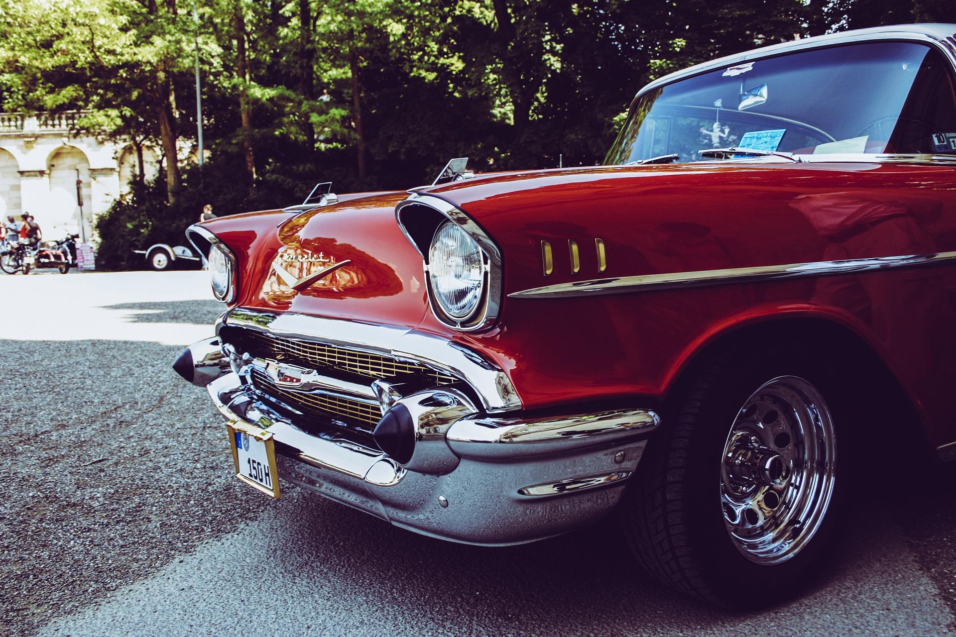 Red classic car, chrome details, parked outdoors near a building and trees.