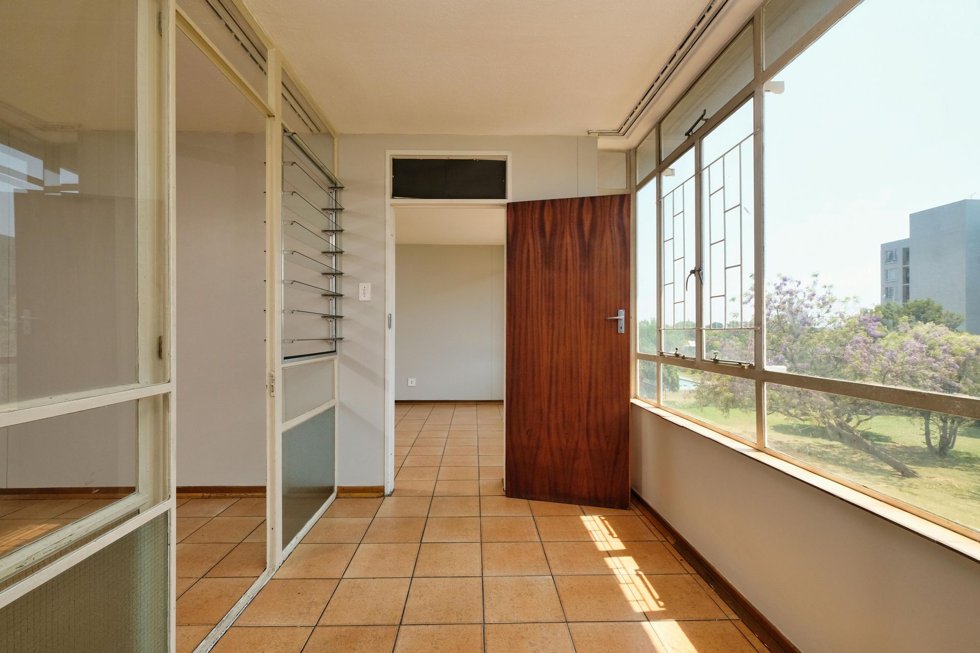 Interior view of a balcony hallway with tiled floor, glass windows, and a wooden door leading to an interior room.