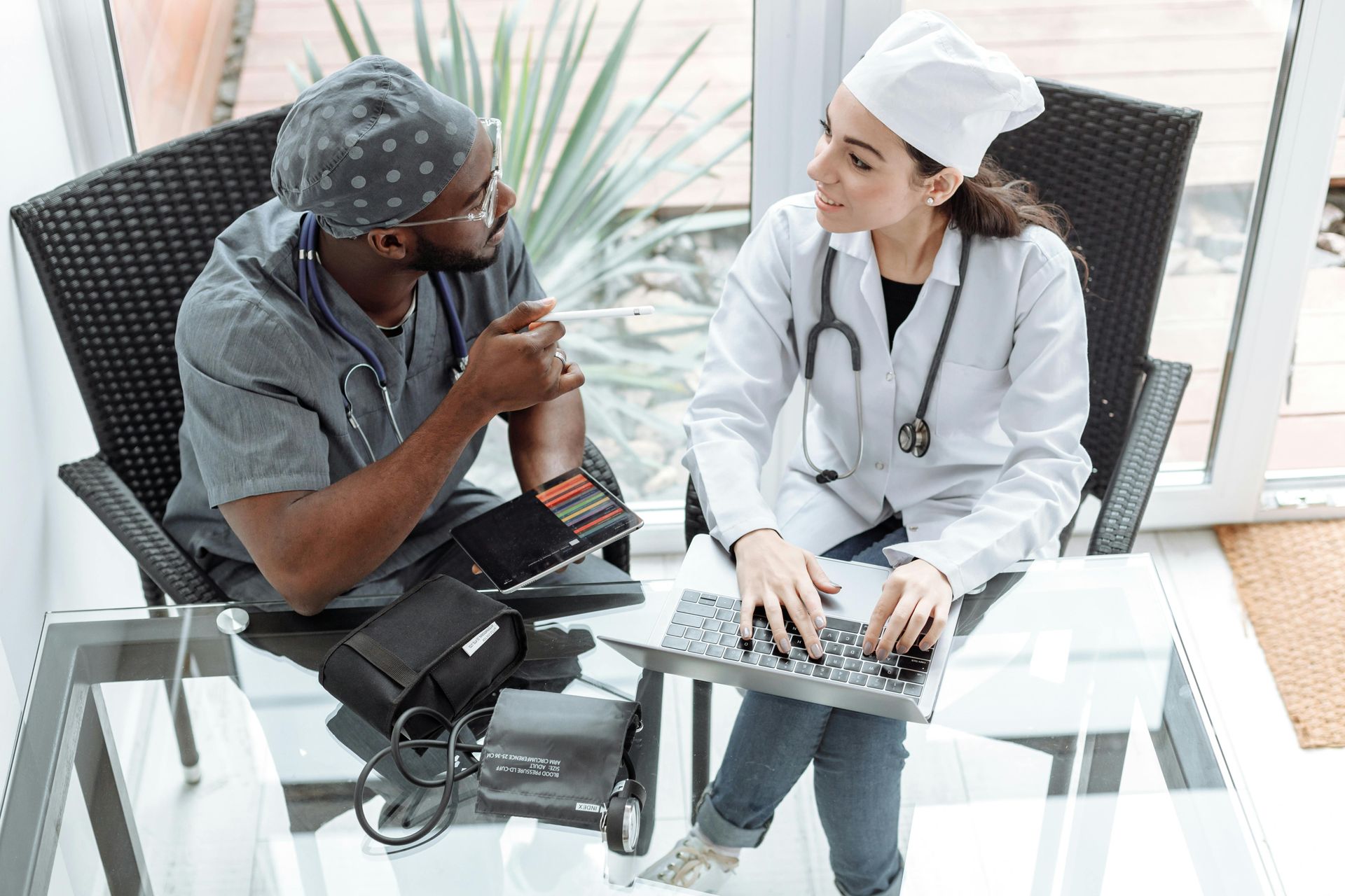 Two medical professionals in scrubs, discussing, with medical equipment on glass table.