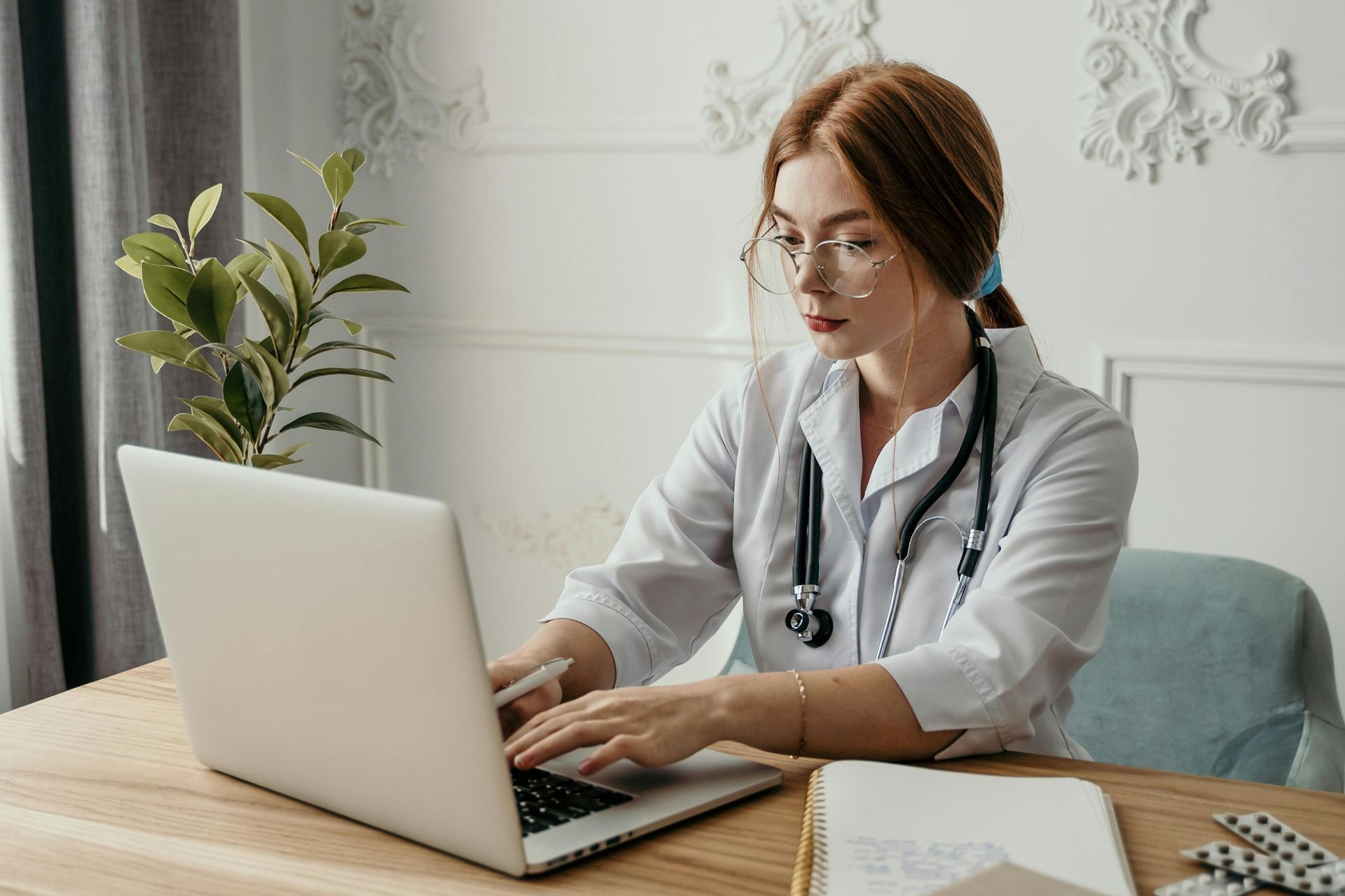 Woman in a white lab coat with a stethoscope typing on a laptop in a well-lit room.