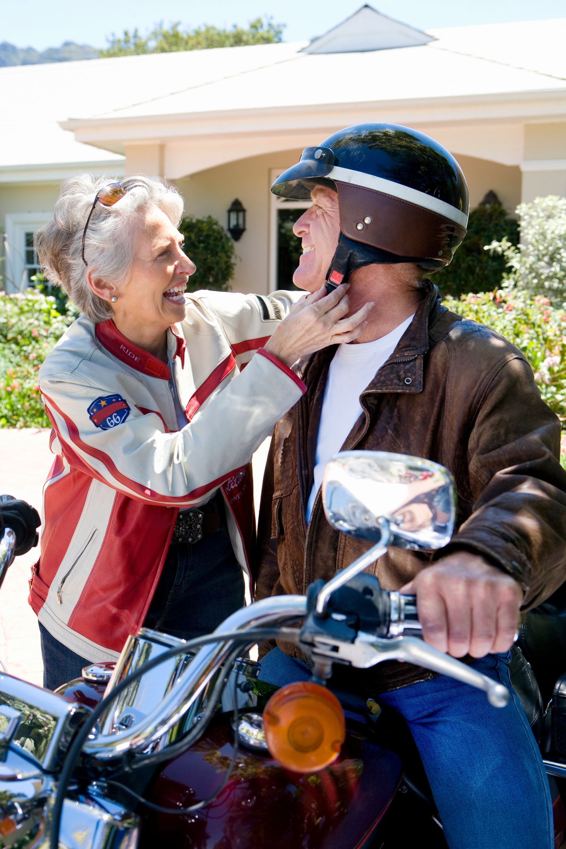 Woman adjusting man's motorcycle helmet outdoors, near house. They are smiling.