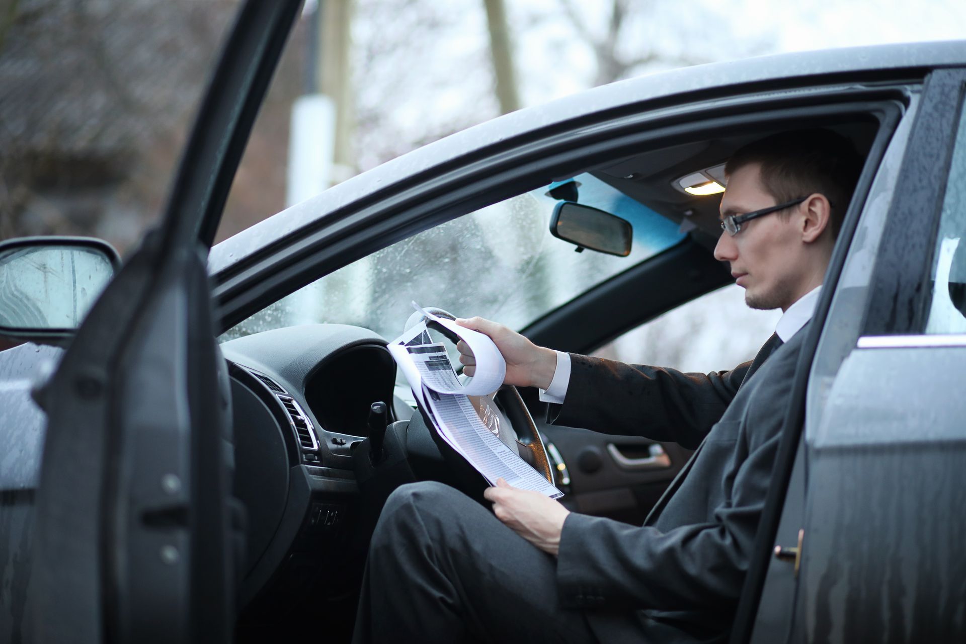Man in suit sitting in car, reading papers. Car door open, focus on documents.