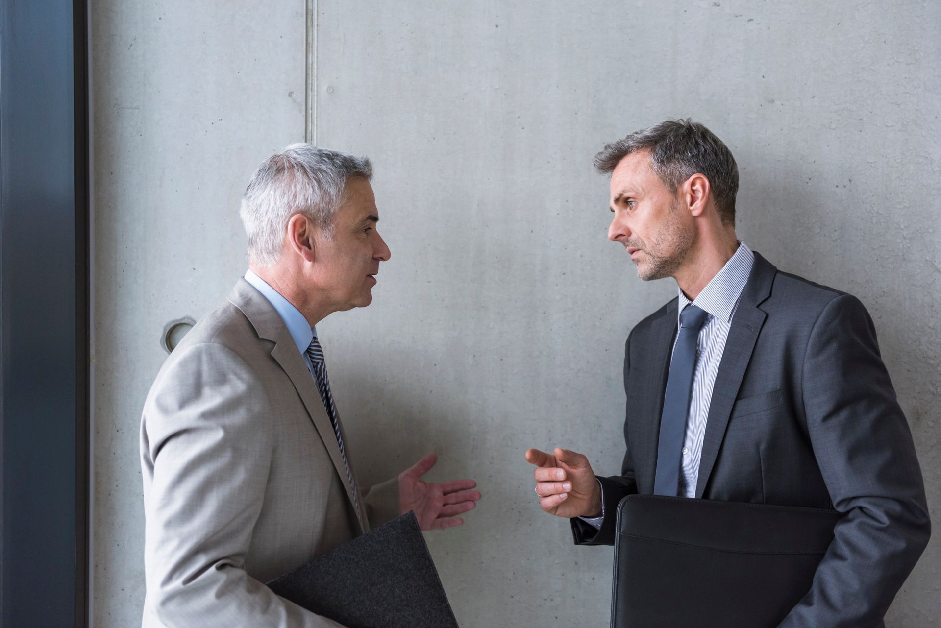 Two men in suits, conversing intently near a concrete wall; one gesturing, the other holding a briefcase, appears focused.