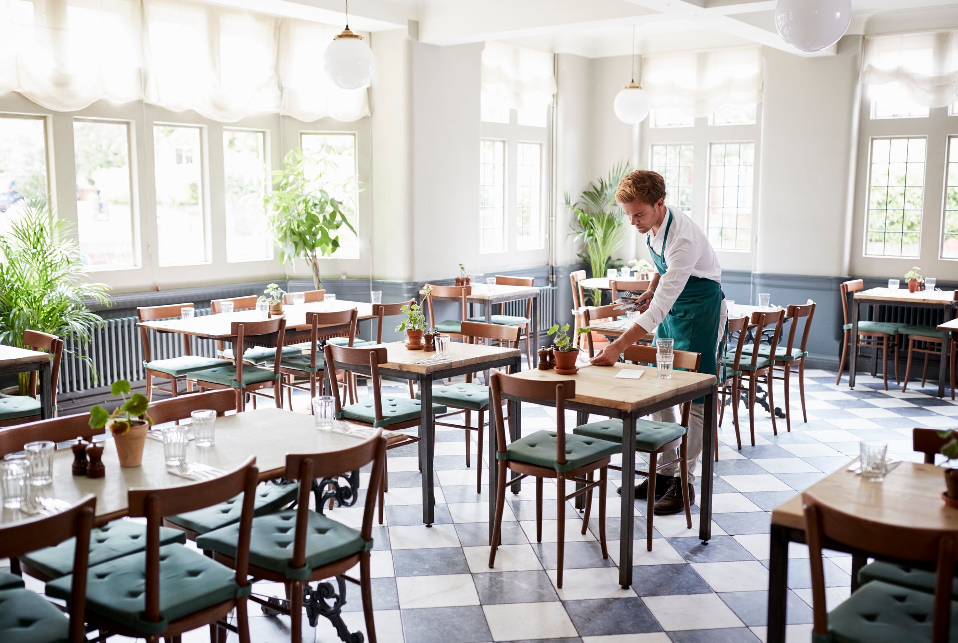 Waiter setting tables in a bright restaurant with checkered floor and large windows.