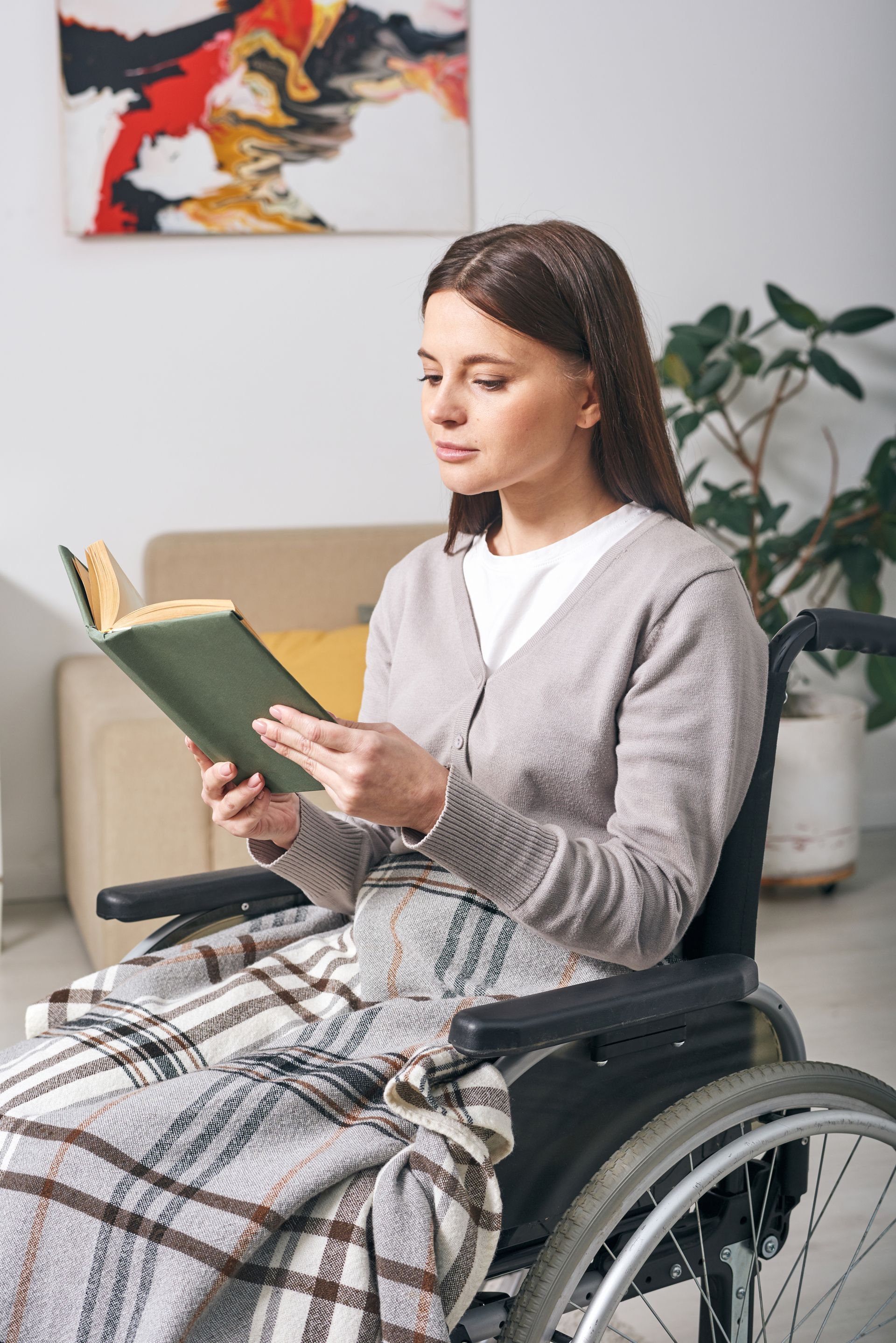 Woman in wheelchair reading a book, wrapped in a blanket indoors.