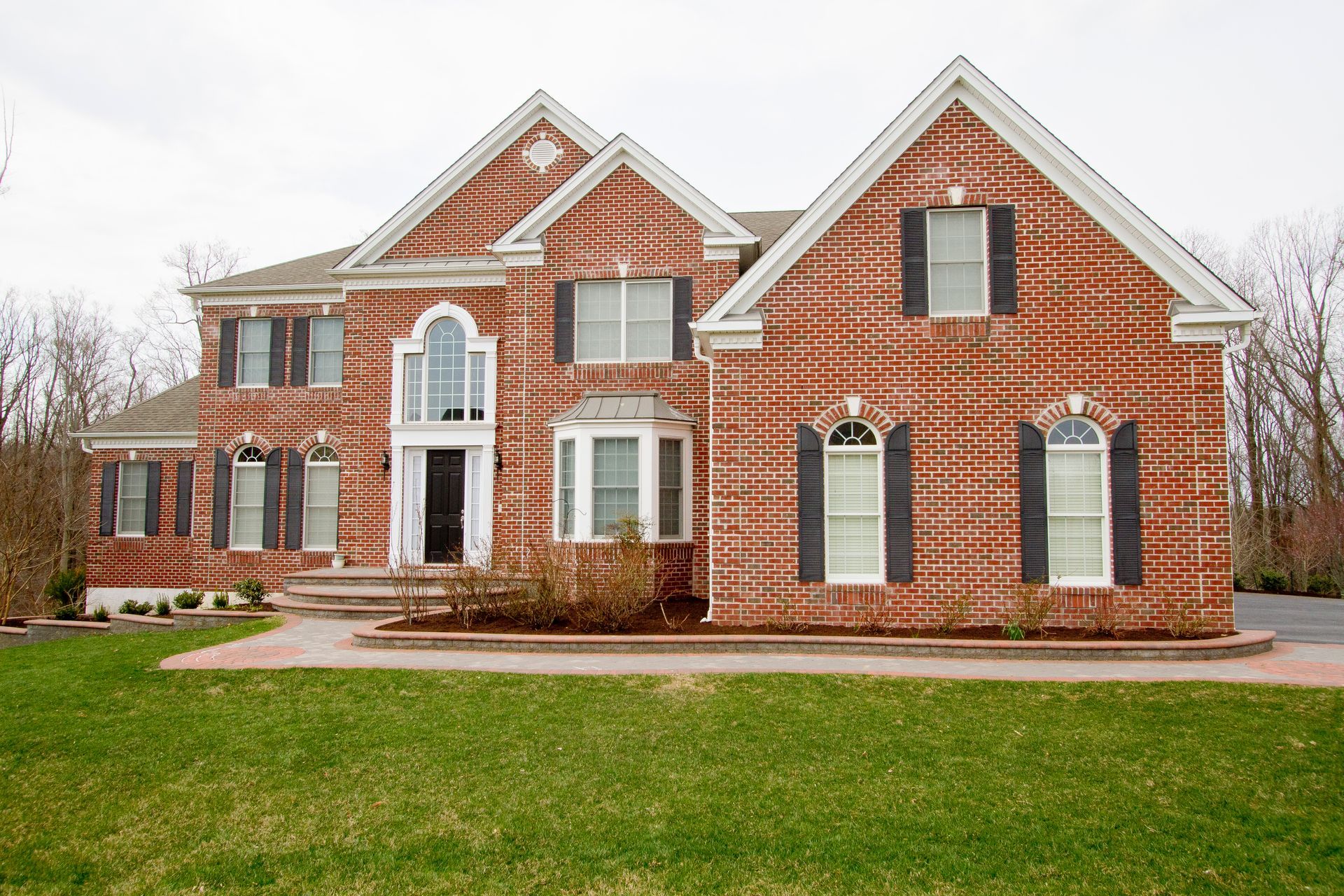 Two-story red brick house with black shutters, green lawn, and a cloudy sky.
