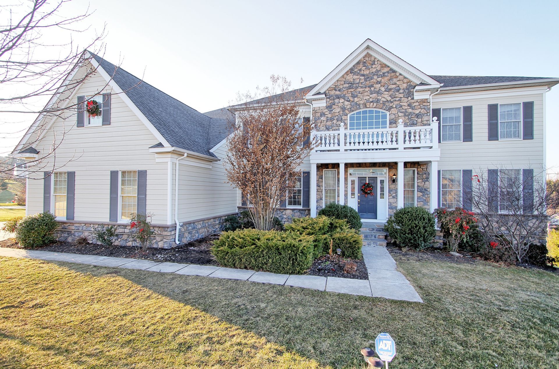 Two-story beige house with stone facade, balcony, shutters, and Christmas wreath; sunny day.