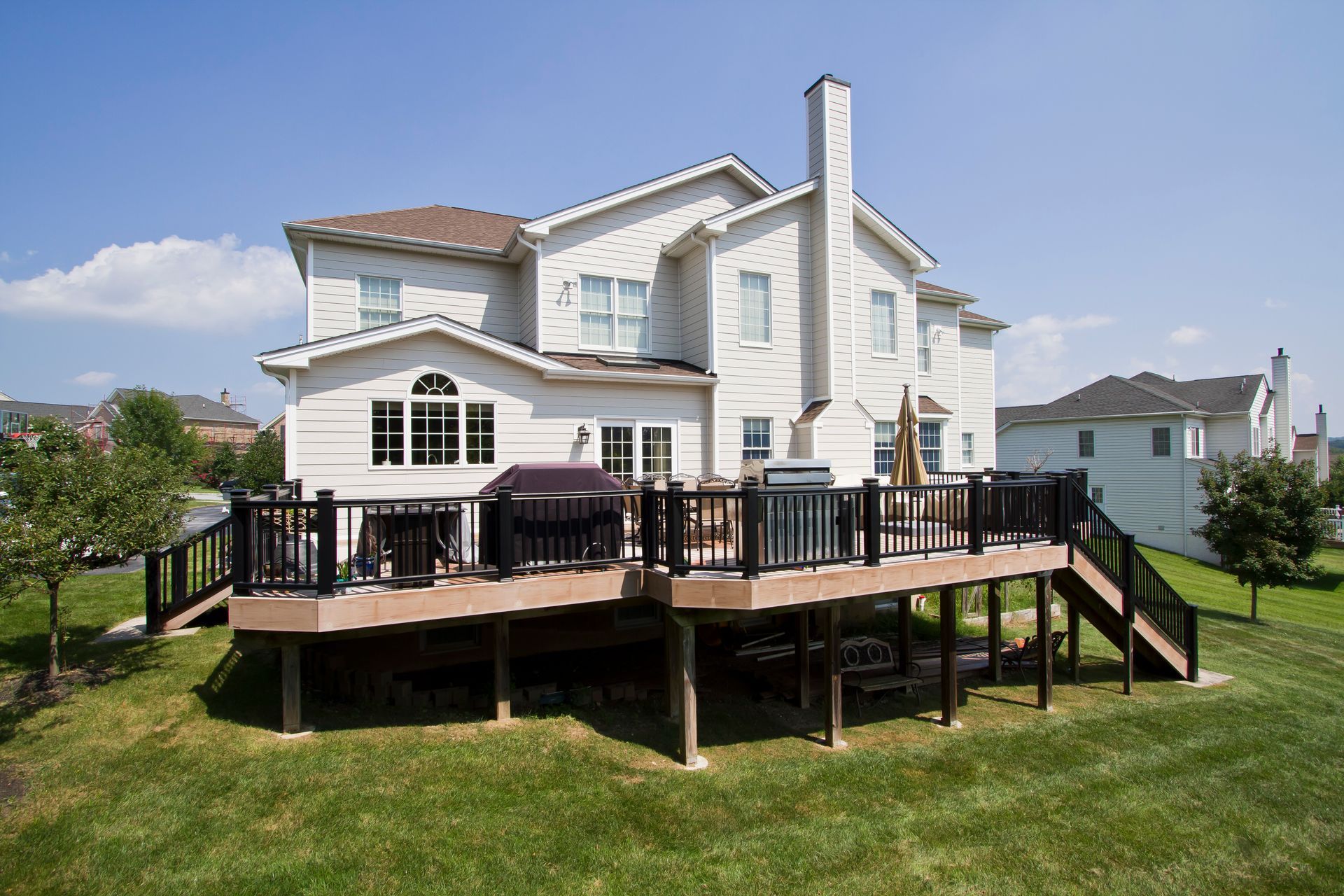 Two-story beige house with a large wooden deck and a green lawn on a sunny day.