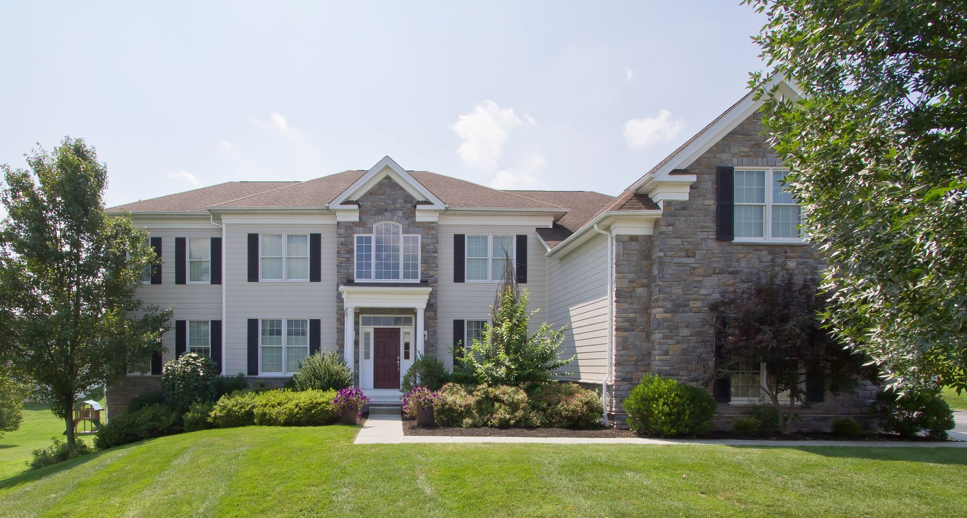 Two-story beige house with stone accents, black shutters, red front door, and manicured lawn.