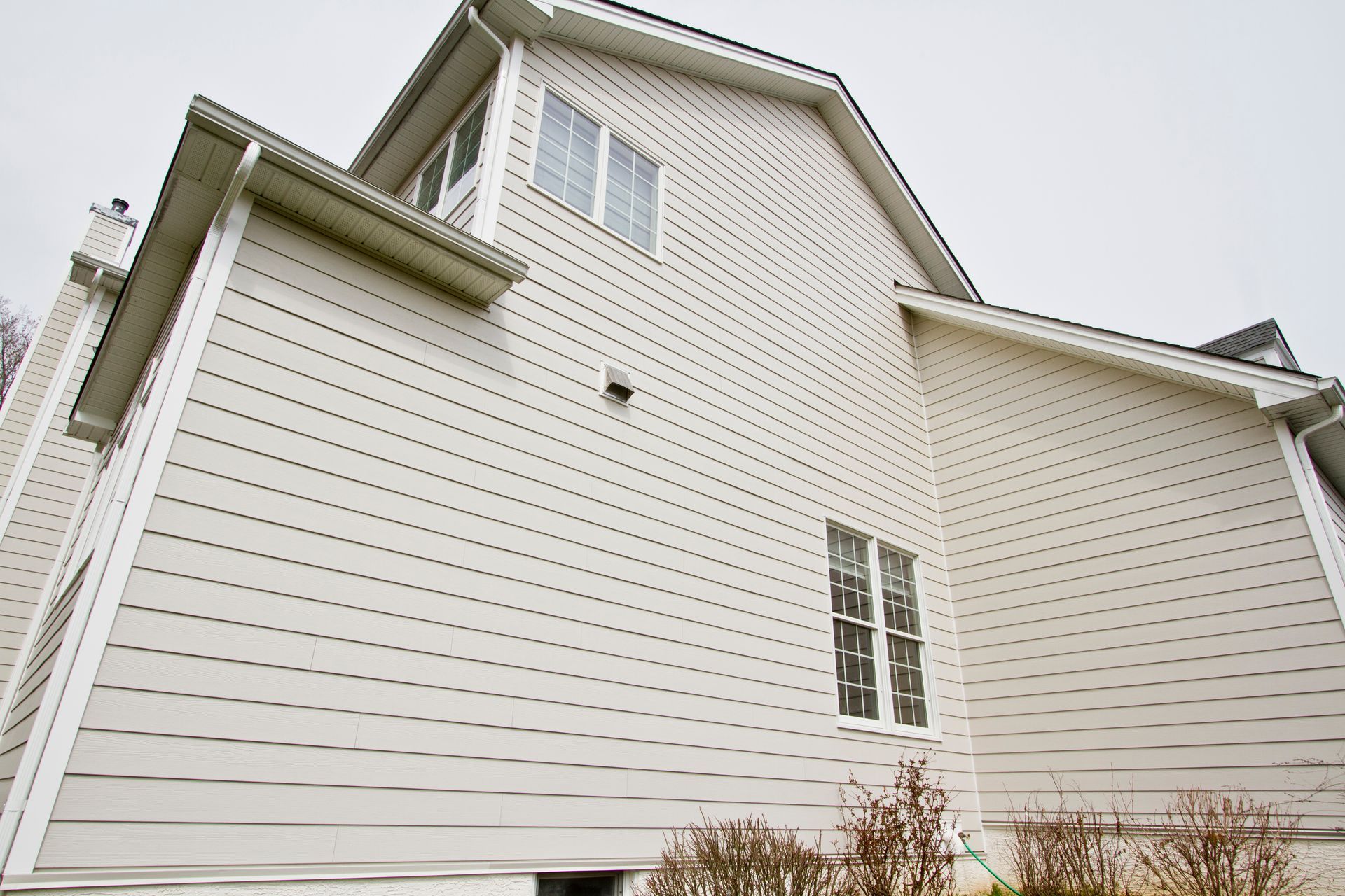 Beige siding on a house with two windows and a vent, under an overcast sky.