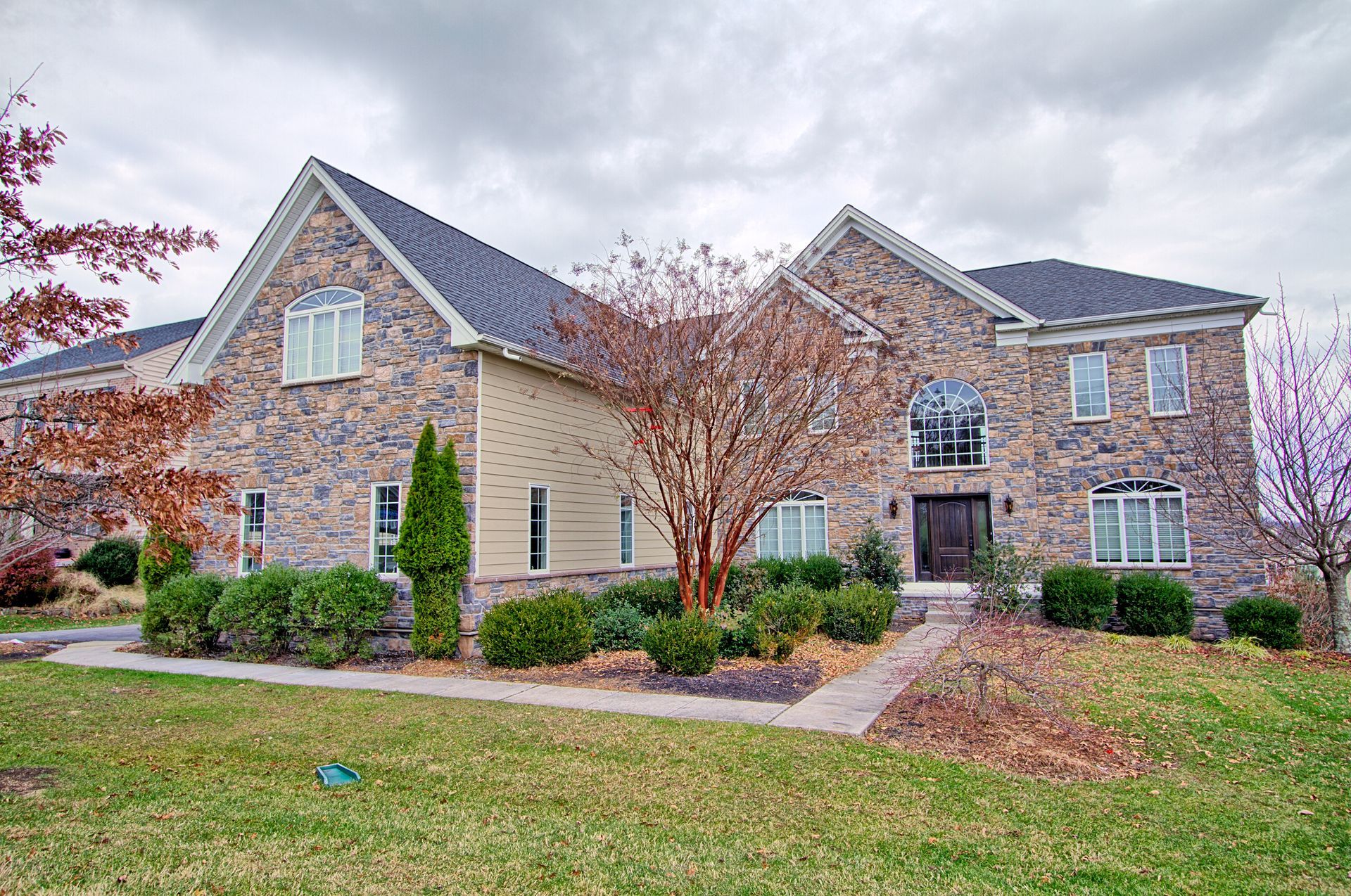 Stone-faced house with dark roof and manicured lawn under a cloudy sky.