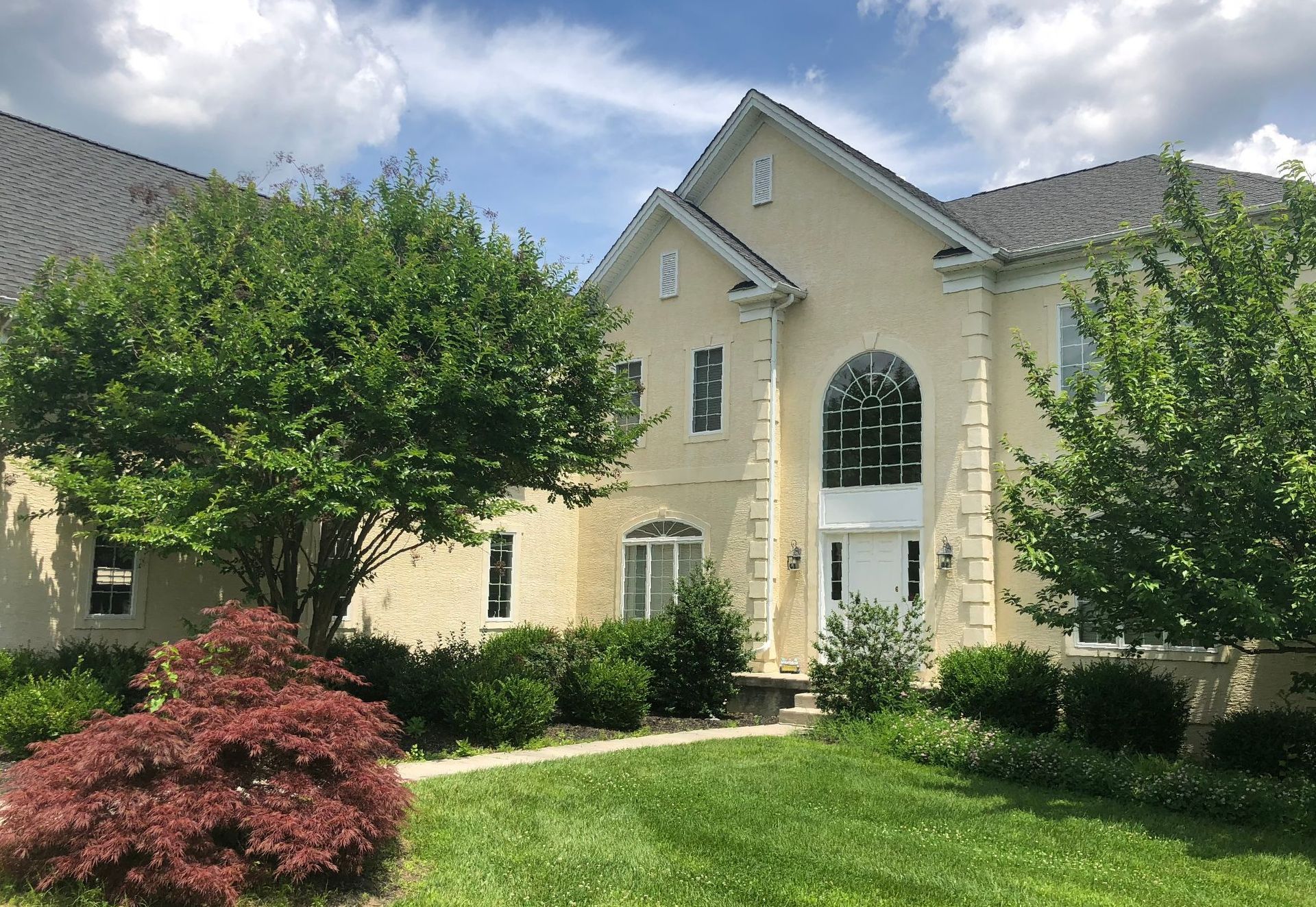 Beige two-story house with gray roof, arched windows, and lush green and red bushes in front.