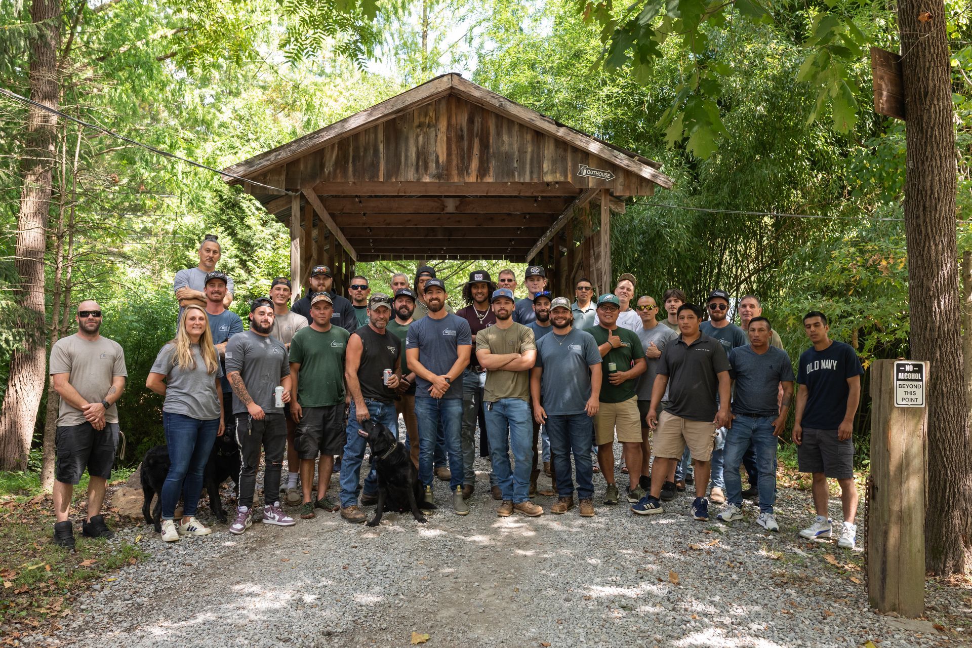 A large group poses for a photo standing on a gravel path in front of a rustic wooden covered bridge surrounded by trees.