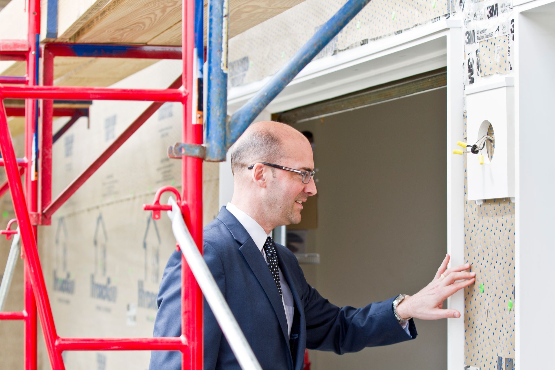 Man in suit inspecting a construction site doorframe, with scaffolding and exposed wall visible.
