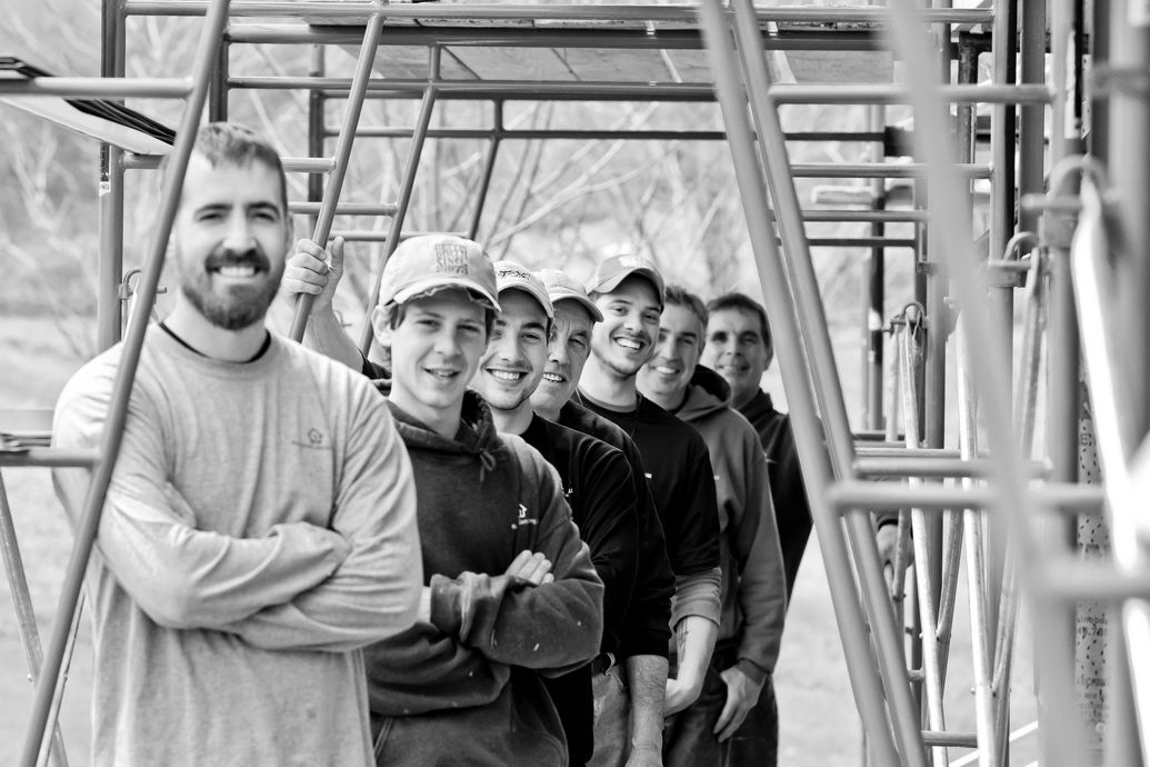 Group of construction workers posing with arms crossed, smiling. Standing near scaffolding.