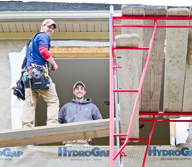 Two construction workers installing a window. One smiles on scaffolding, the other in the window frame.