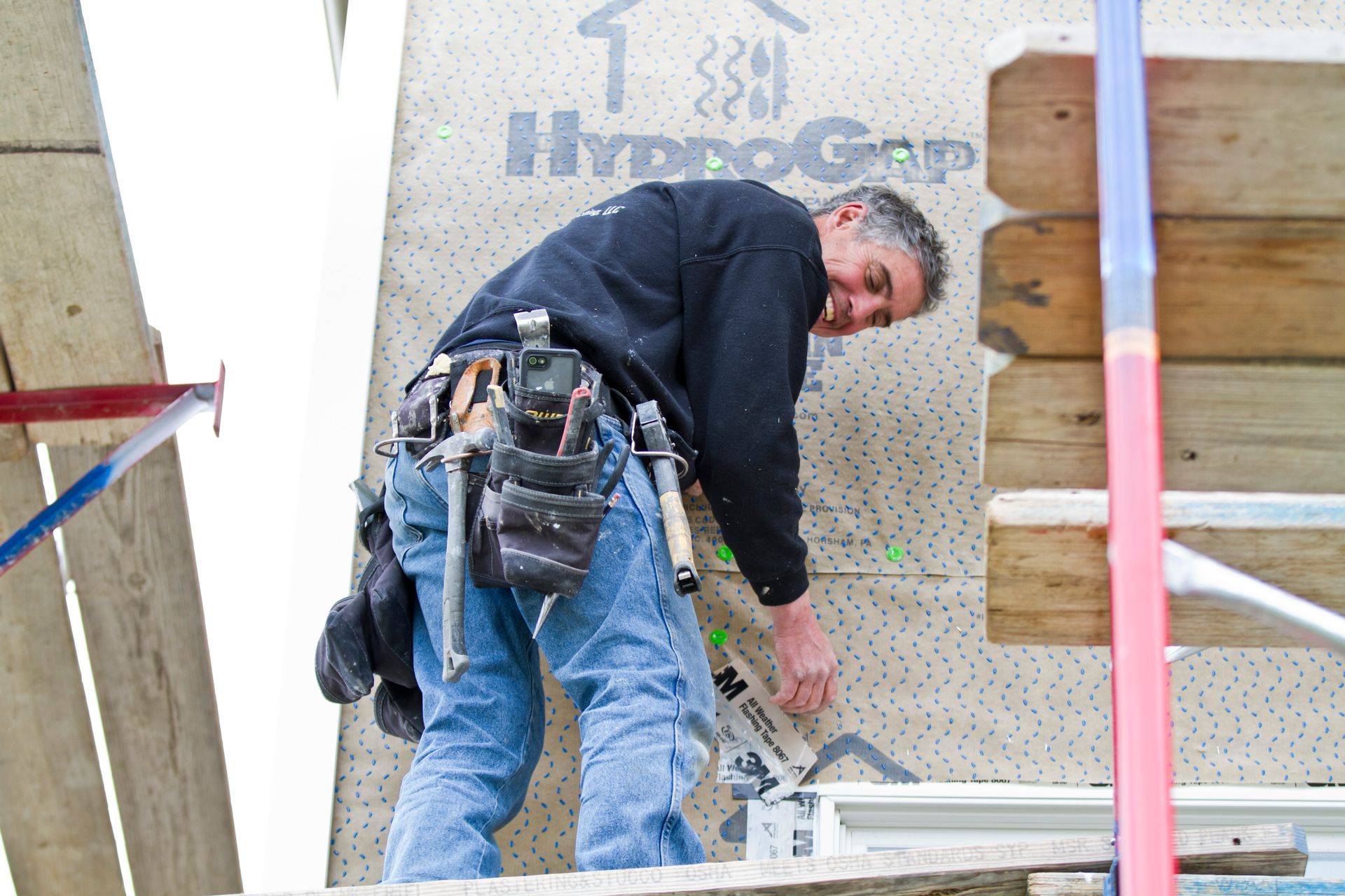 Construction worker in blue jeans and black shirt measuring siding on a building.