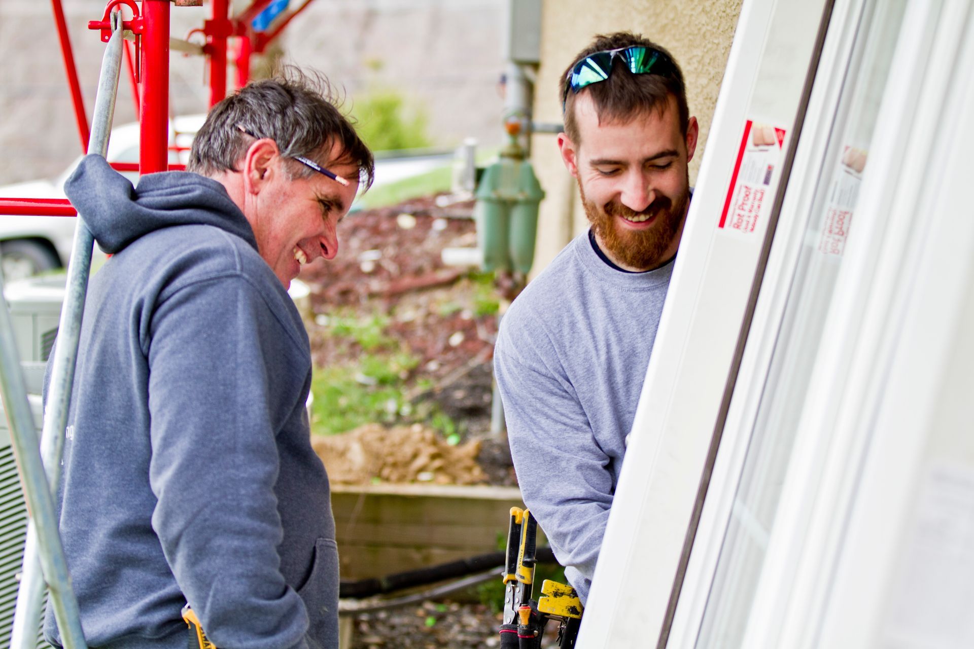 Two construction workers, outdoors, smiling as they handle a large white door.