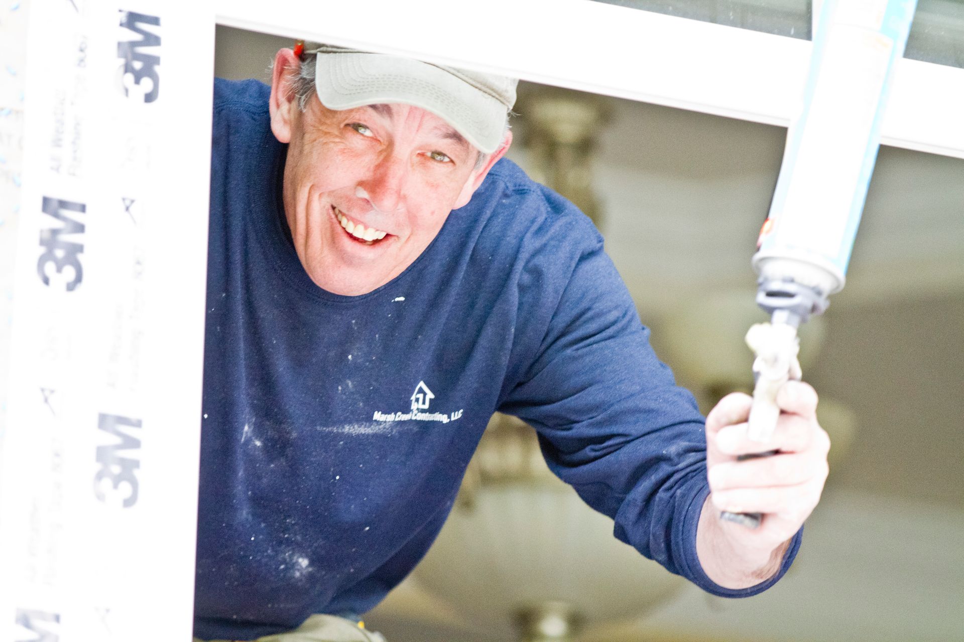Man with cap, applying foam sealant around a window frame, smiling.