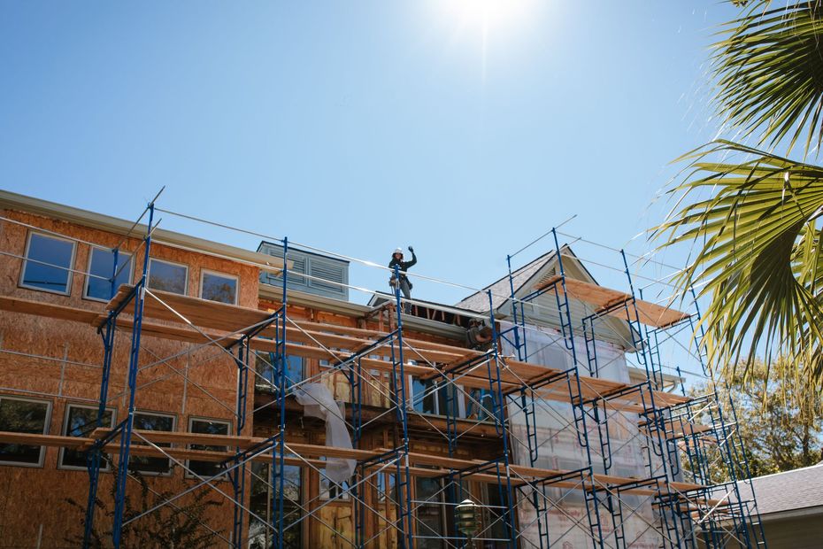 Construction of a two-story building with scaffolding and a worker on top under a bright blue sky.