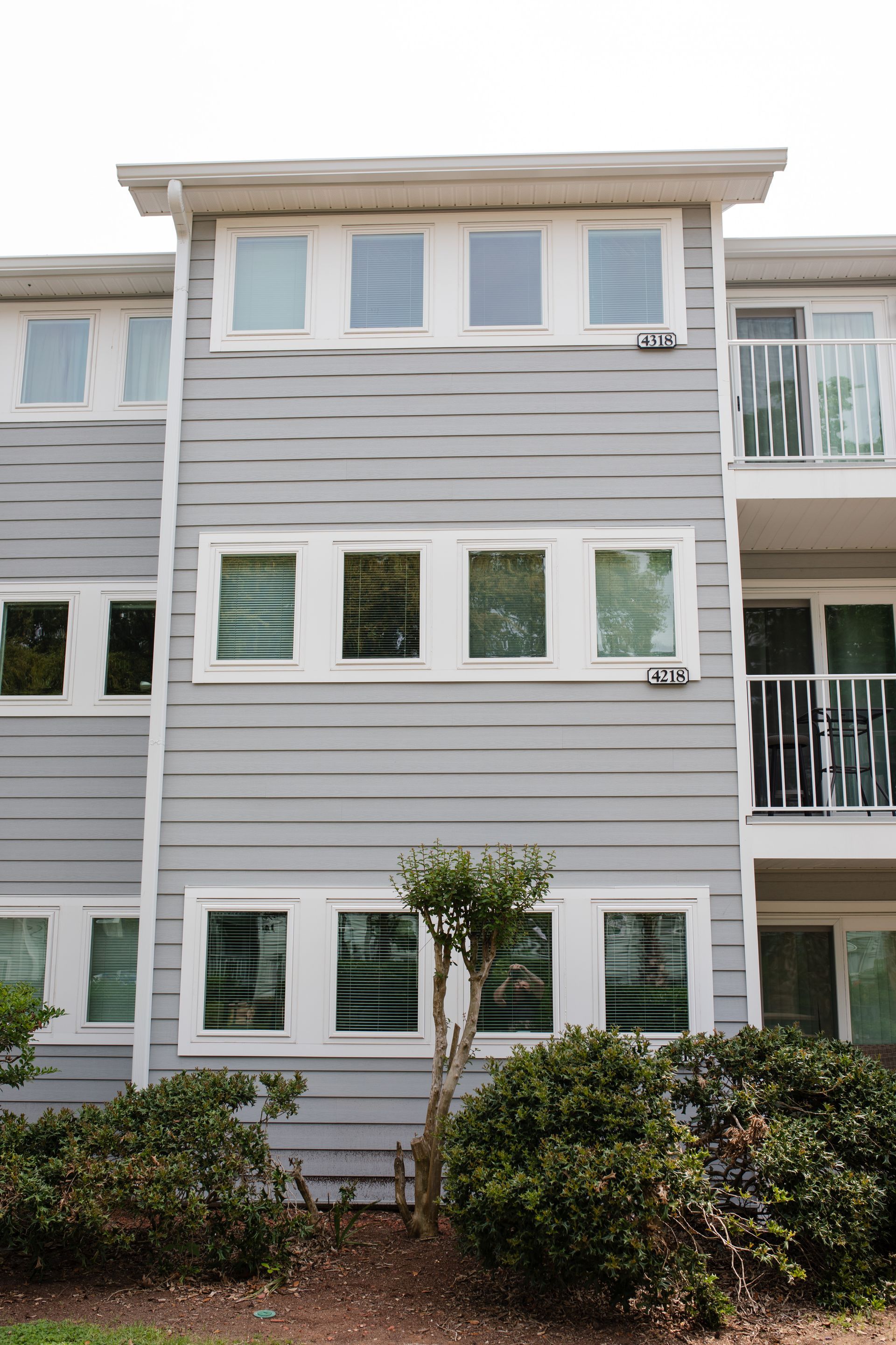 Gray apartment building with white-framed windows and a small balcony. Bushes and a tree are in the foreground.