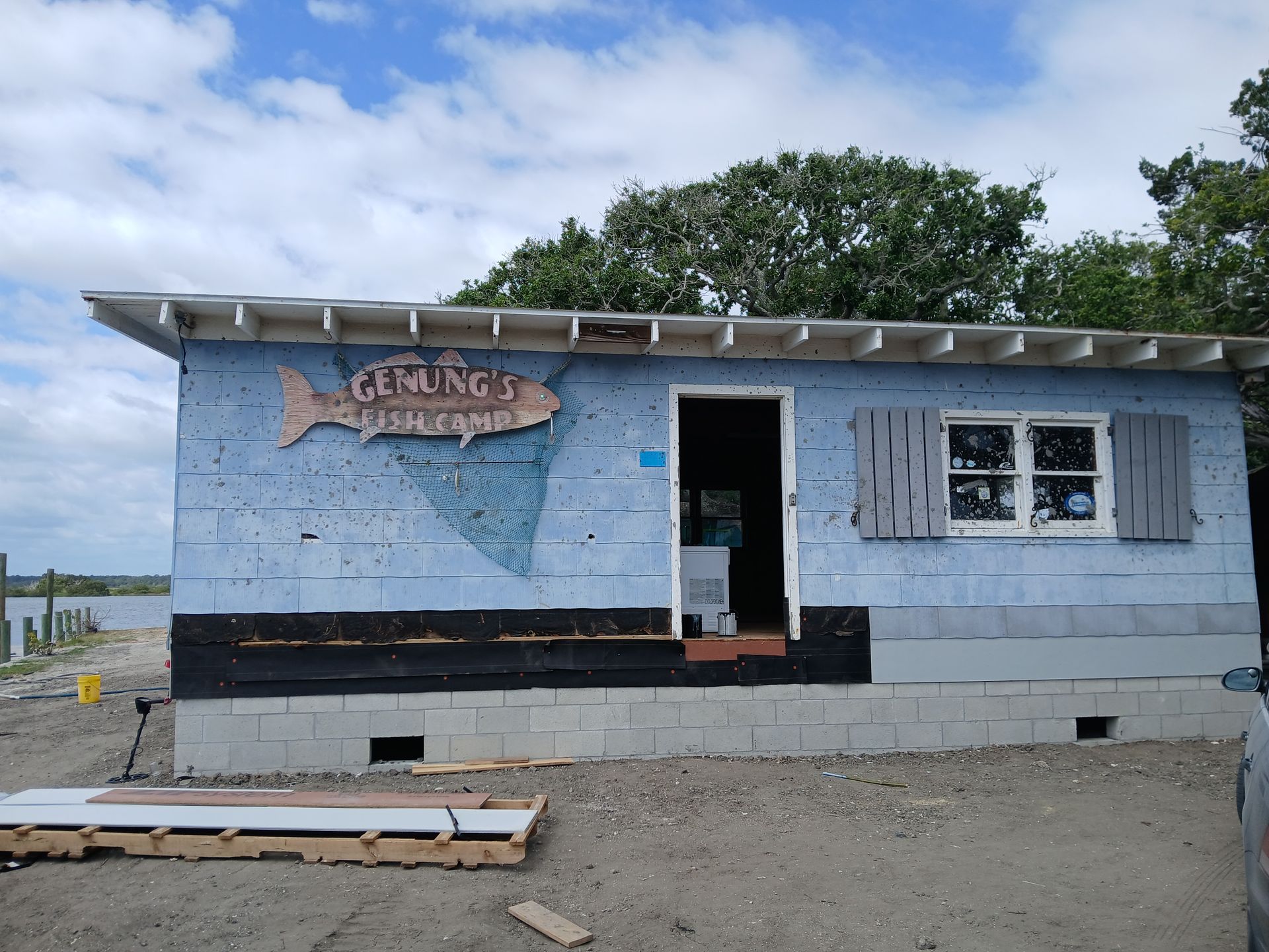 Small blue beach shack with sign, open door and windows, on a sandy shore under a cloudy sky.