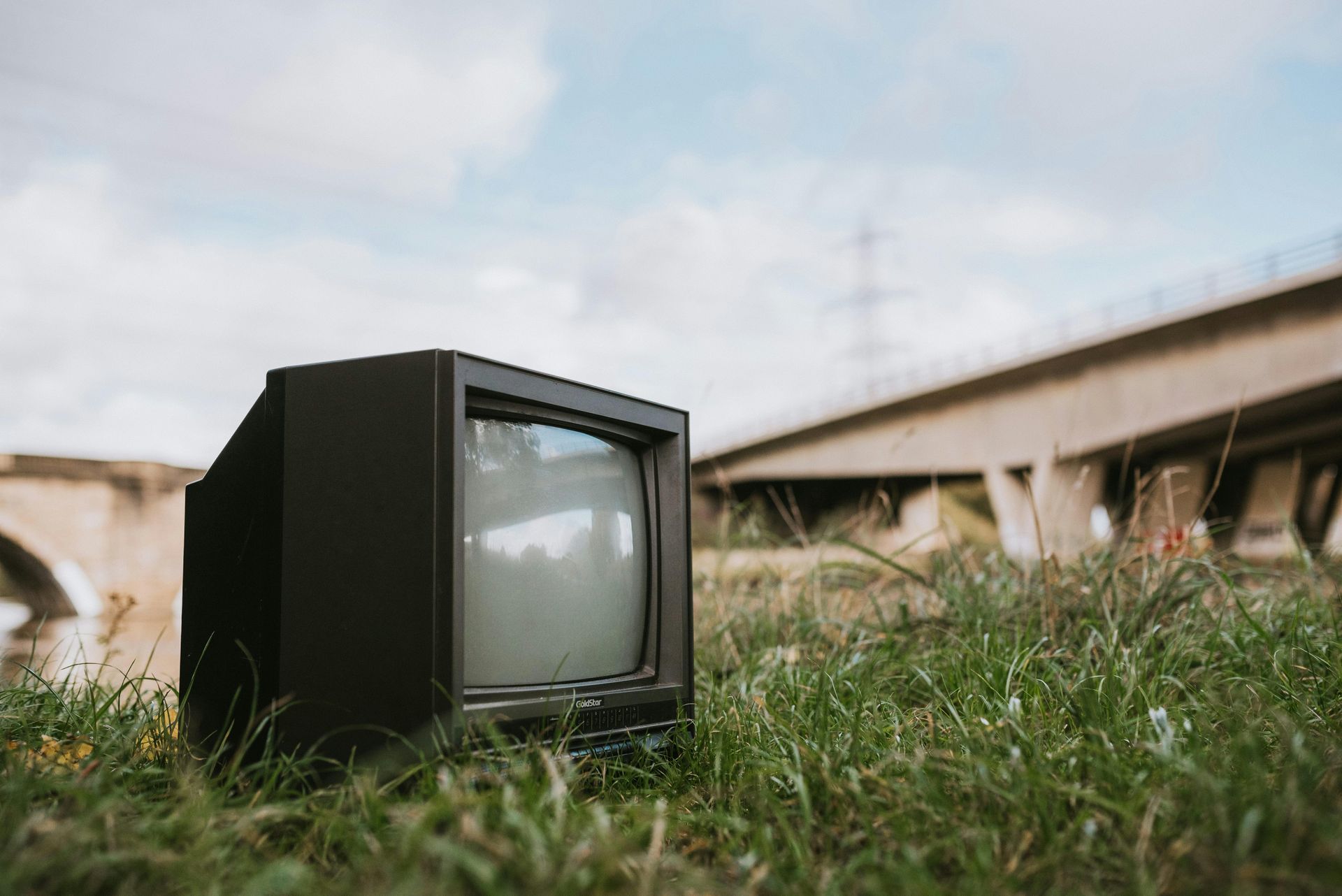 An old television is sitting in the grass in front of a bridge.