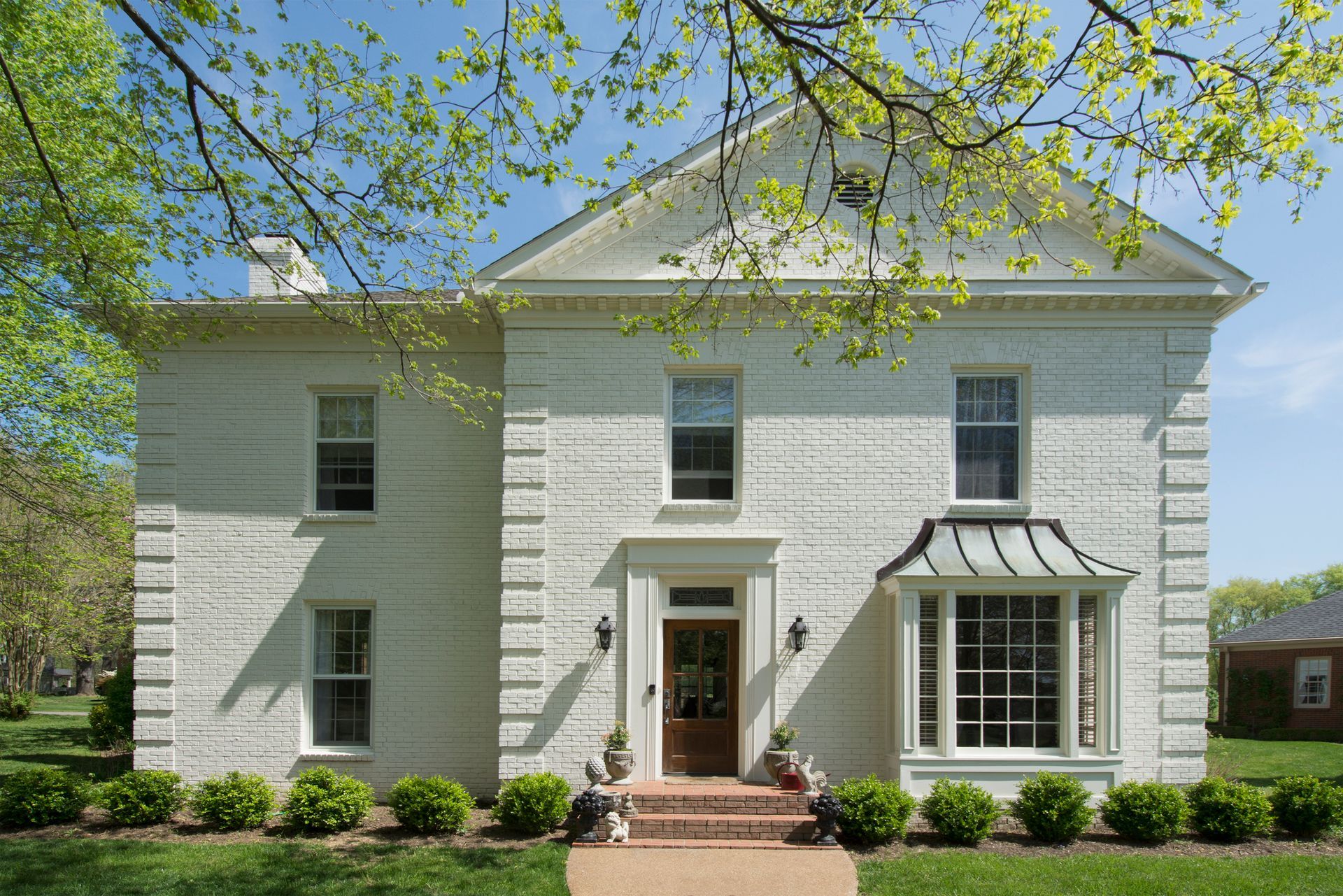 White brick house with symmetrical design, bay window, and arched doorway. Lush green lawn and trees surround the home.