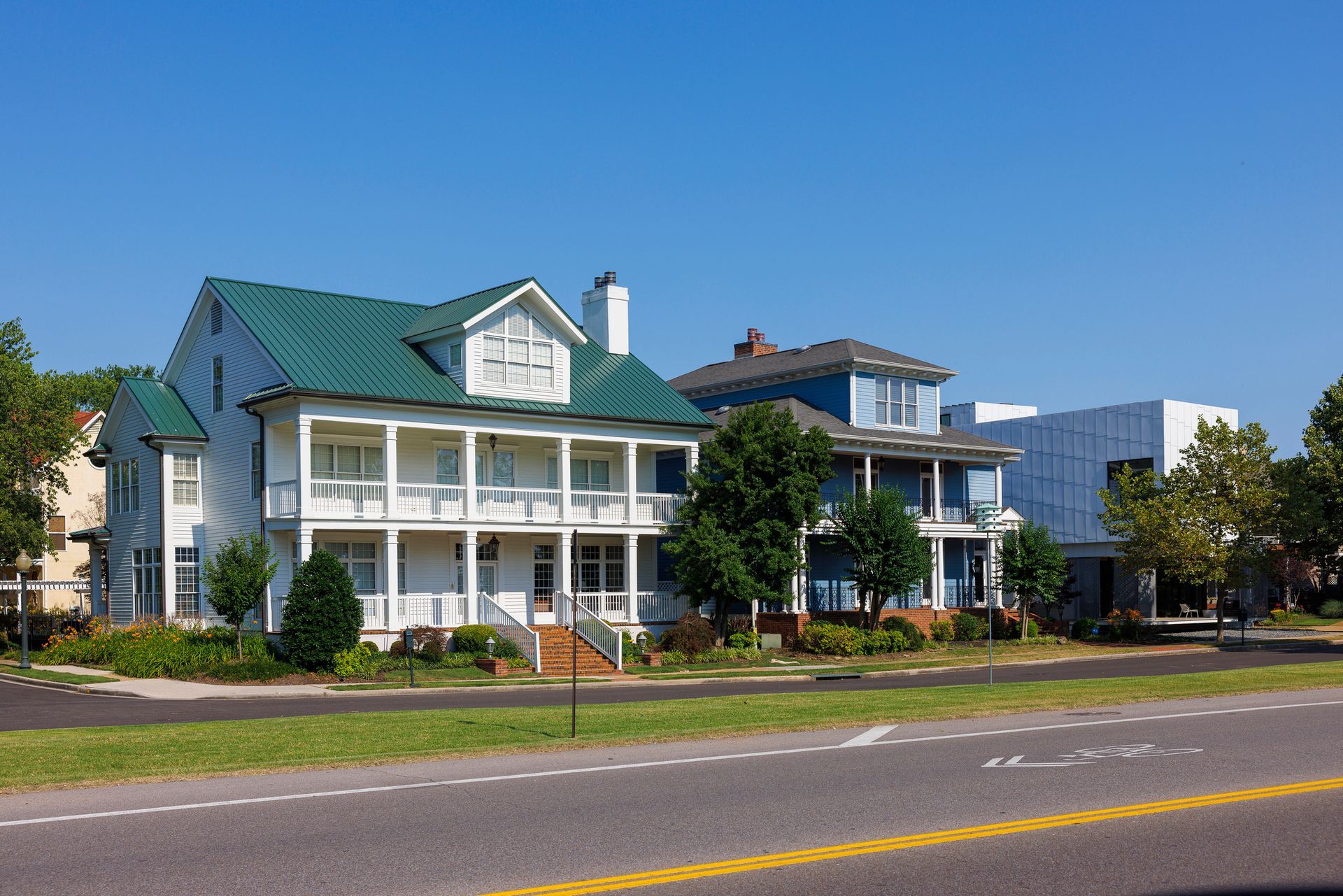 Two-story white house with green roof, a blue house, and a modern white building next to a road, under a clear sky.