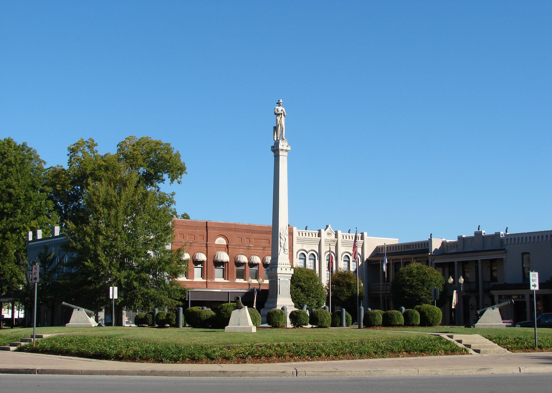 Monument in a grassy square with surrounding buildings under a clear blue sky.